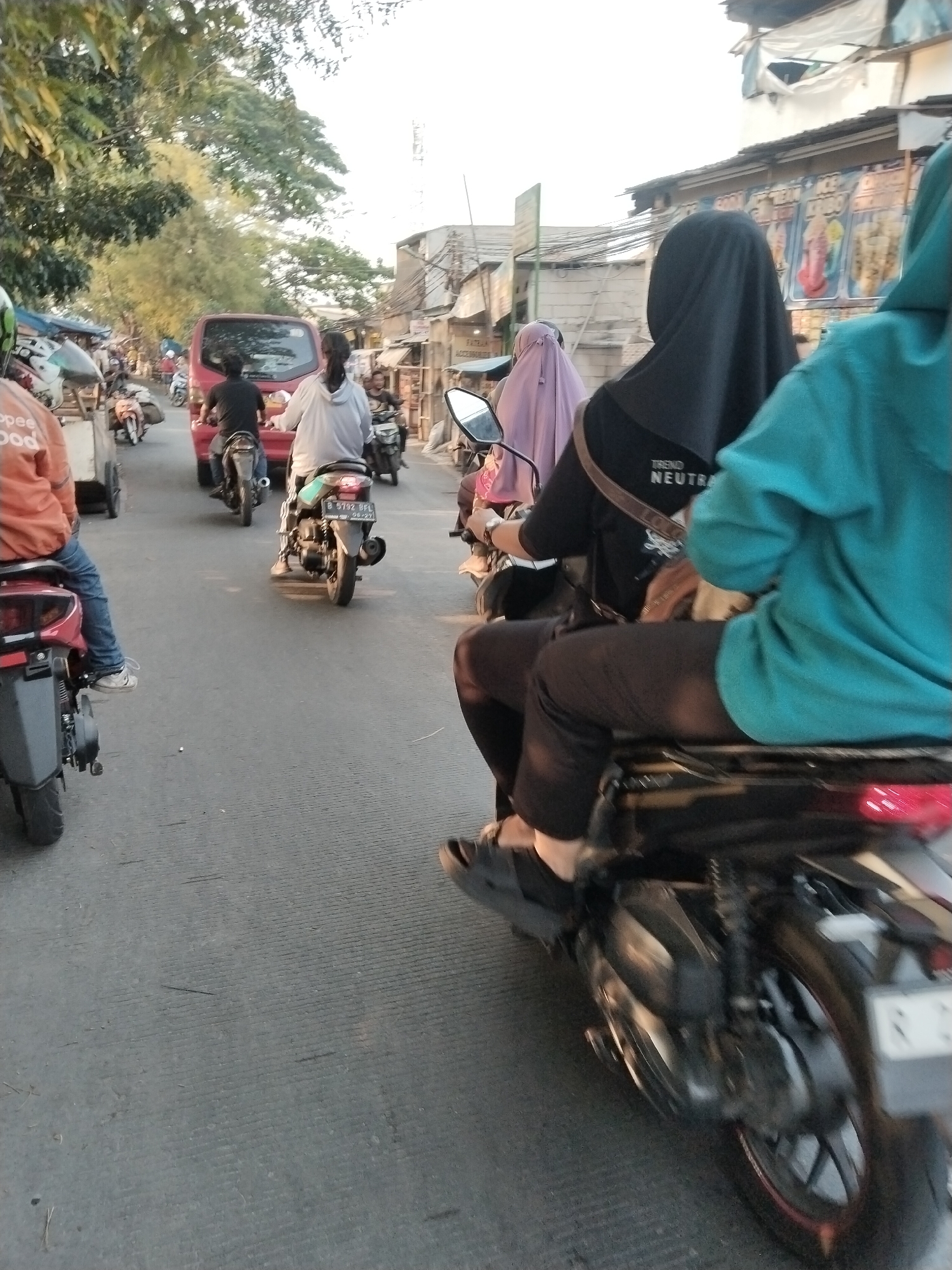 A street scene in West Jakarta, Indonesia, shows a traffic jam with numerous motorbikes and a red car. The perspective is from the back of a motorbike, looking forward. In the foreground, the rider of the motorbike in front is wearing an orange shirt and blue jeans. To the right, another motorbike rider is wearing a teal jacket and black pants, and a person behind them is wearing a black headscarf and shirt. Further ahead, a person in a light-colored hoodie is on a motorbike behind the red car. The red car has a license plate that is partially visible. Buildings and trees line the street, with some appearing to be shops. The sky is bright, suggesting daytime. The road surface is paved and appears somewhat worn.