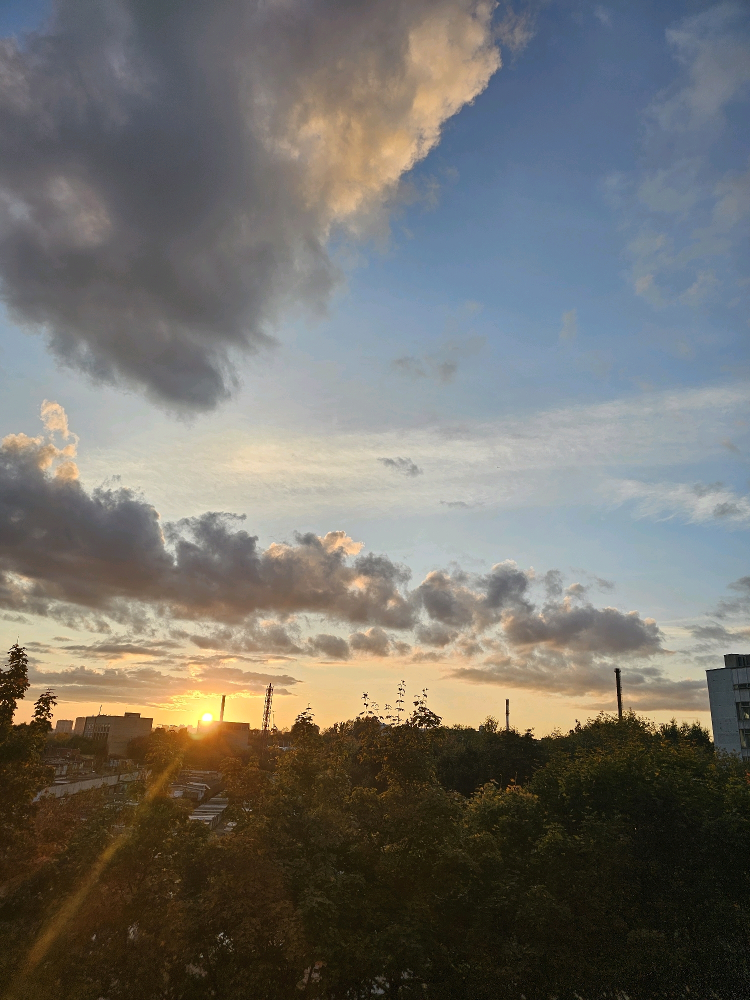 The image depicts a cityscape at sunset, with the sun low on the horizon and casting warm light. The sky is filled with a mix of dark and bright clouds, with patches of blue sky visible. In the foreground, there is a dense canopy of green trees. Beyond the trees, the silhouette of buildings and industrial structures can be seen. Two tall, dark smokestacks rise above the treeline in the middle ground. A light-colored building with windows is visible on the right side of the frame. The scene appears to be an urban environment. The location is identified as Khimki, Russia. There are no visible people or animals in the image, nor any apparent actions or interactions.