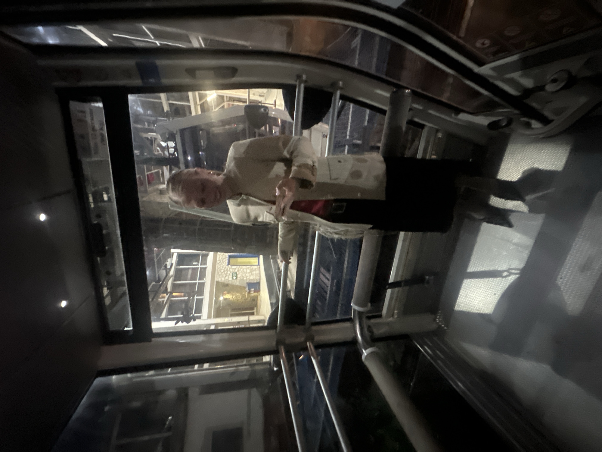 The original image, rotated 90 degrees counter-clockwise, captures an interior view from what appears to be a cable car cabin at night.
**Subjects:**
A person with short, light-colored hair is visible in the reflection of the glass window on the left side of the frame. They are wearing a light-colored jacket or shirt over a red garment and dark trousers or a skirt, looking towards the camera with a neutral to slightly smiling expression, one hand seemingly gesturing. The cable car's interior features dark paneling, structural elements, and large windows.
**Scene and Setting:**
The scene is set inside a cable car, likely in motion during the evening or night. Through the windows, illuminated buildings and structures of a town are visible against the dark night sky. The external environment is softly lit by artificial lights from the distant buildings.
**Activity or Event:**
The image depicts a passenger riding in a cable car. The person in the reflection seems to be observing the view or perhaps posing for a photograph.
**Notable Details:**
The time of day is night, emphasized by the dark exterior and reliance on artificial light sources both inside and outside the cable car. On one of the buildings seen outside, a blue and yellow banner or flag, strongly resembling the flag of Ukraine, is visible. The internal lighting of the cable car is dim, creating reflections on the glass. The context provided specifies the location as Taormina, Italy, suggesting this is the local cable car system. No other discernible text is clearly legible in the image or its crops.