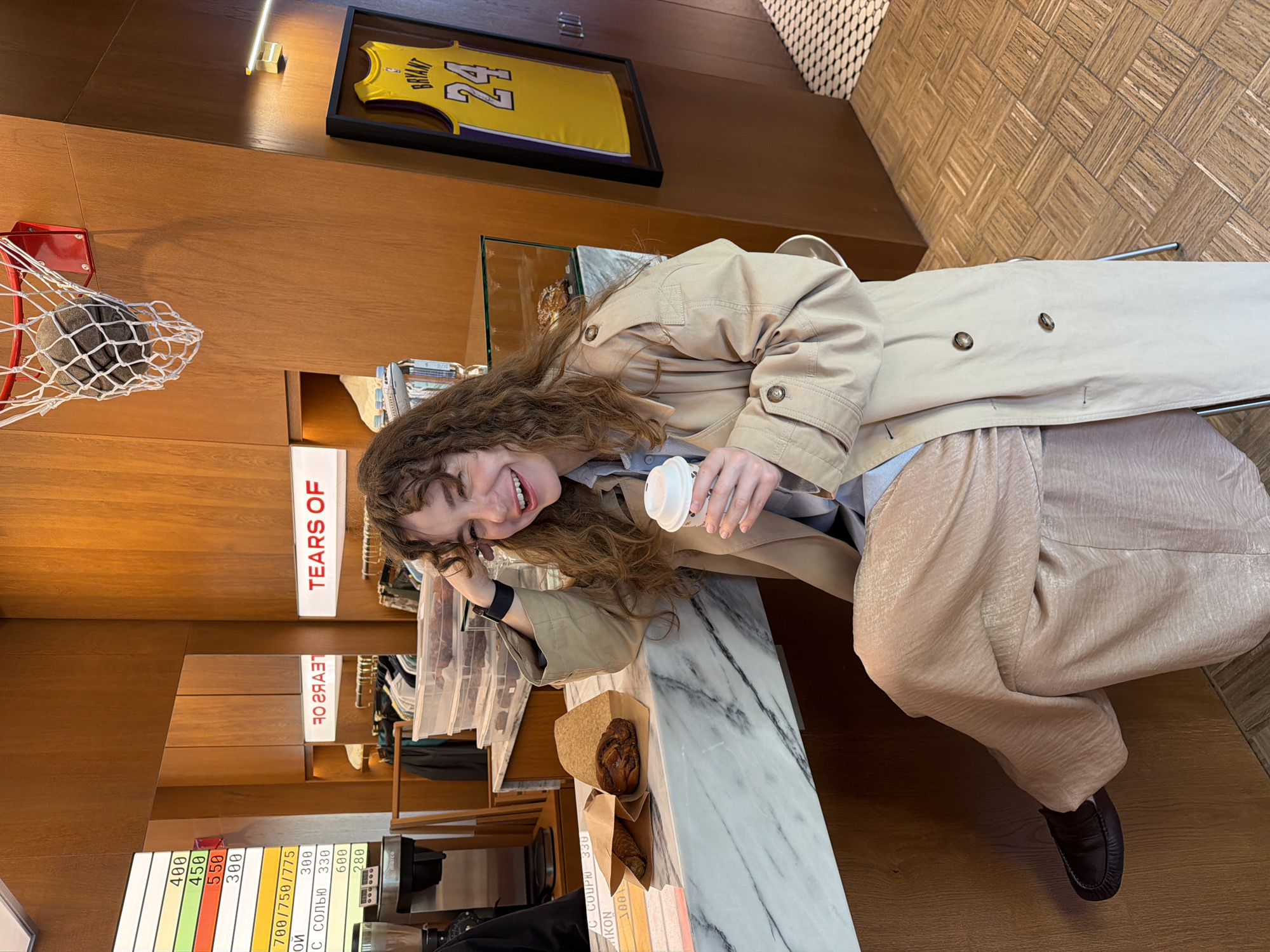 A woman with curly brown hair, wearing a light beige trench coat and flowy, light-colored pants, is captured sitting at a marble-topped counter in what appears to be a modern cafe or shop in Moscow, Russia. She is smiling broadly and holding a white disposable coffee cup in her right hand. Her left hand rests near her face, and a dark watch is visible on her wrist.
The setting features warm wooden walls. Above the counter, a framed yellow basketball jersey with the name "BRYANT" and the number "24" is displayed. To the left, a small red basketball hoop with a net holds a gray, textured basketball. A light fixture runs horizontally above the jersey.
On the counter next to the woman, two pastries in brown paper bags are visible. Behind her, a lighted sign reads "TEARS OF". Further to the left, a menu board displays various items with prices in what appears to be rubles. Visible text on the menu includes "400", "450", "550", "300", "700/750/775", "ОЙ", "300", "ОГАТ", "с солью 330", "600", "280", "СОУРЮ 330", "IKON", and "100". Another small white sign reads "marco". The floor has a patterned wooden tile design. The overall impression is one of leisure and enjoyment in a stylish, well-lit indoor environment during the daytime.