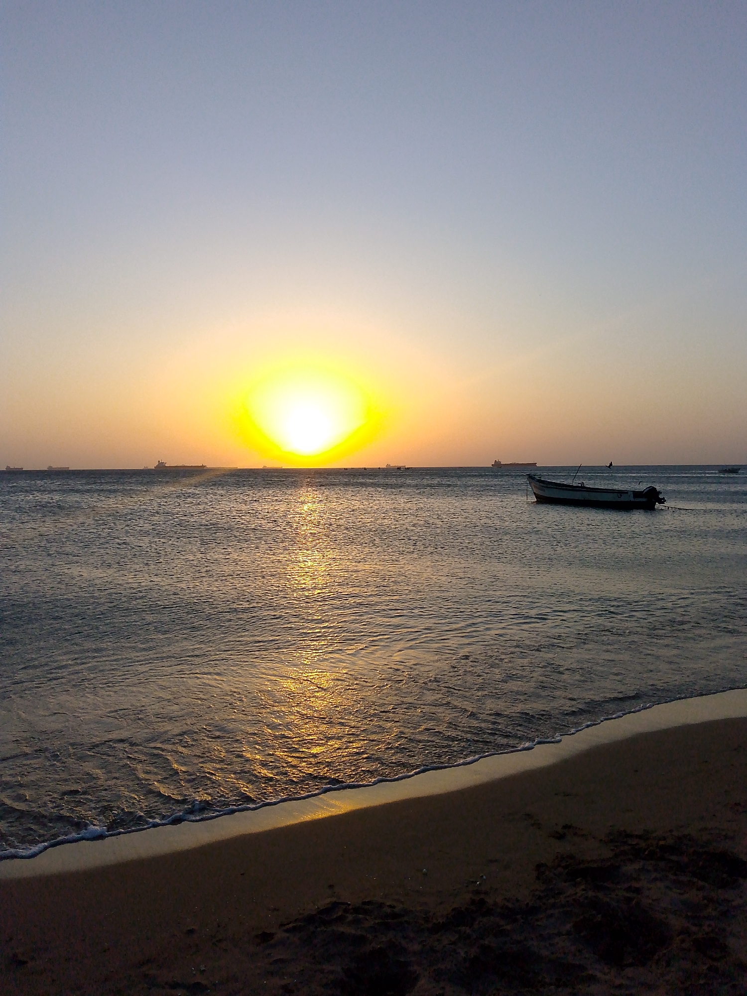 The image depicts a sunset scene at Villa Marina, Venezuela. A body of water extends from the horizon to the bottom of the frame, with sunlight reflecting off its surface. A boat with a motor is positioned in the water to the right. Several ships are visible along the horizon. The foreground includes a sandy beach with gentle waves. The sky transitions from light blue to yellow, with the sun centered on the horizon. There are no people visible in the image.