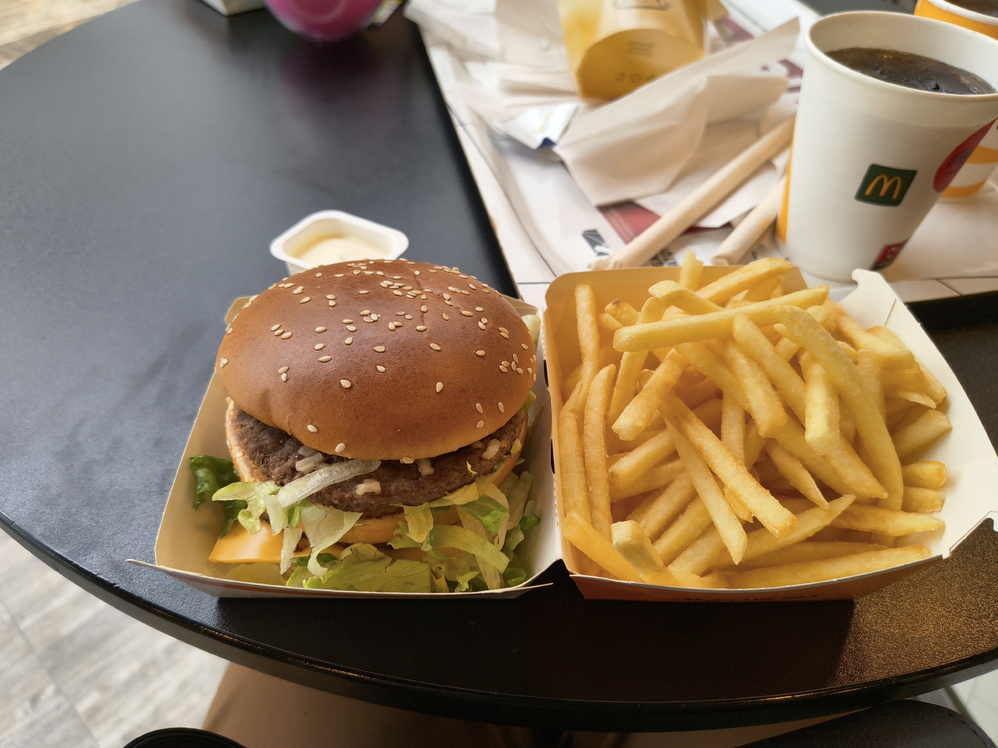 A meal is displayed on a dark, reflective surface, likely a table. In the foreground, a burger in a cardboard container sits to the left, with a side of french fries in a separate cardboard container to the right. The burger appears to be a Big Mac, with three layers of bun, two beef patties, lettuce, cheese, and onions. The french fries are golden brown and appear to be freshly made. In the background, a white paper cup with a McDonald's logo is visible, containing a dark beverage. Another similar cup, partially obscured, is also present. Several paper straws and a white paper wrapper are scattered on a tray behind the food and drink. The overall lighting suggests an indoor setting, possibly a fast-food restaurant. No individuals are visible in the image.