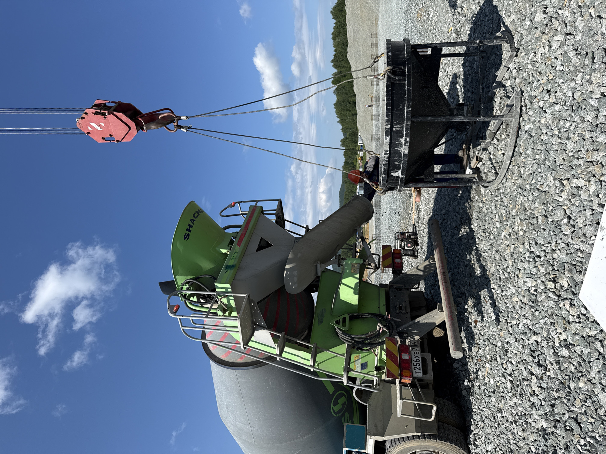 A construction worker in a red hard hat and dark work clothes stands near a large concrete mixer truck. The mixer truck's chute is positioned over a large, black, funnel-shaped concrete bucket, which is suspended by cables from a red crane hook. The scene appears to be a construction site with a gravel-covered ground and a clear blue sky with some clouds. In the background, there are piles of gravel and a treeline, suggesting an outdoor, possibly rural or suburban, location. A license plate on the truck reads "T256XE 74". The location is identified as Leninsk, Russia.