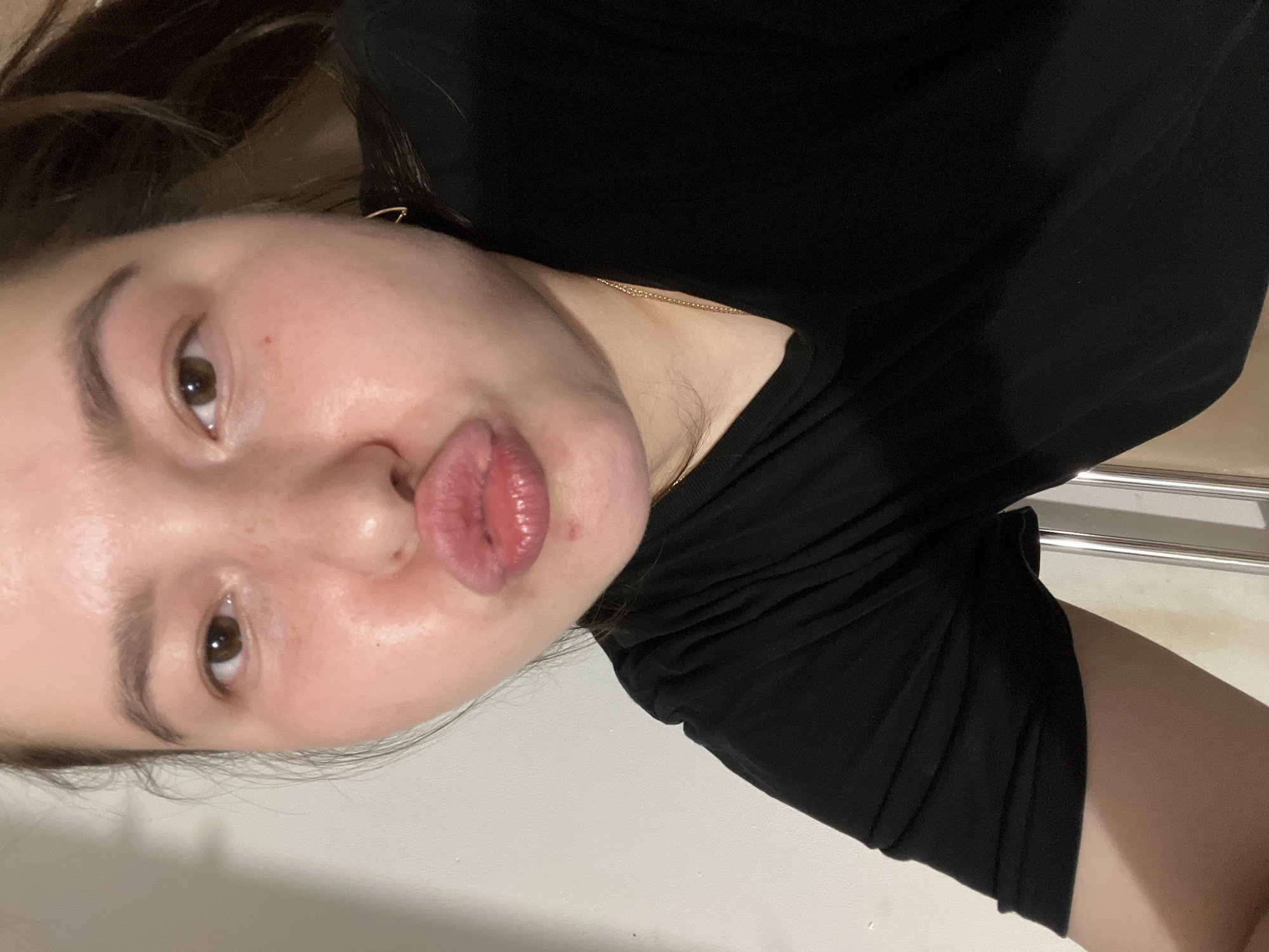 A person with fair skin, brown eyes, and dark hair is shown in a close-up, selfie-style photograph. They are wearing a black t-shirt and a delicate gold chain necklace. The person is making a kissing gesture with their lips and has their tongue slightly extended. The background is a plain, light-colored wall, with a hint of a metal railing visible on the left side. The image is oriented sideways.