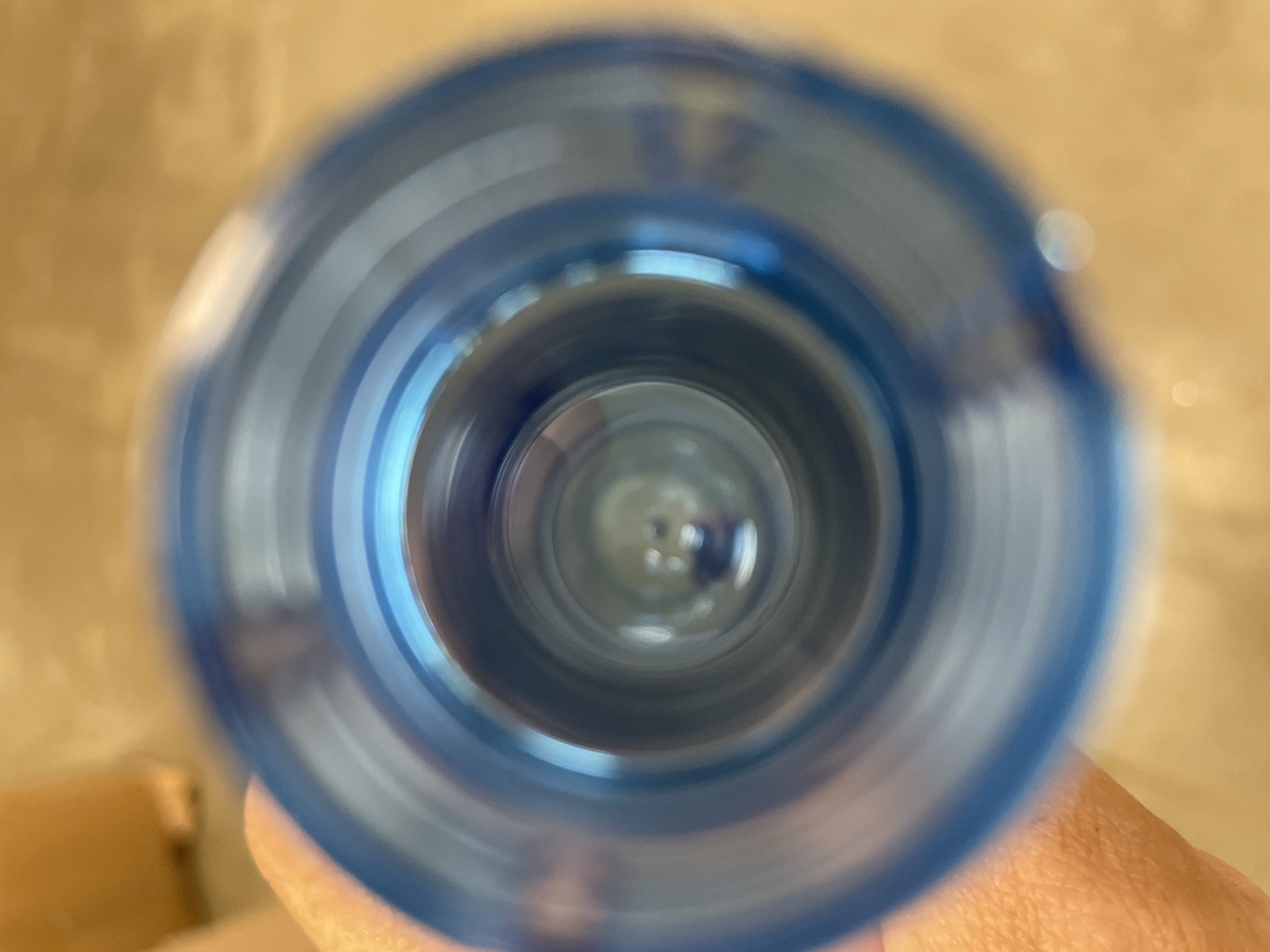 The image shows a close-up, distorted view looking down into the opening of a blue plastic bottle. The perspective is from the viewpoint of someone holding the bottle, with a portion of their hand visible on the left side of the frame, out of focus. The background, visible around the bottle opening and through it, is a blurred, warm-toned surface, possibly a wooden floor or wall. Concentric rings of blue plastic are visible as the sides of the bottle's opening. The center of the opening is a dark, blurry circular area, with faint reflections suggesting a shiny inner surface or liquid. The scene does not contain any identifiable landmarks or elements that would specifically place it in Терволово, Russia.