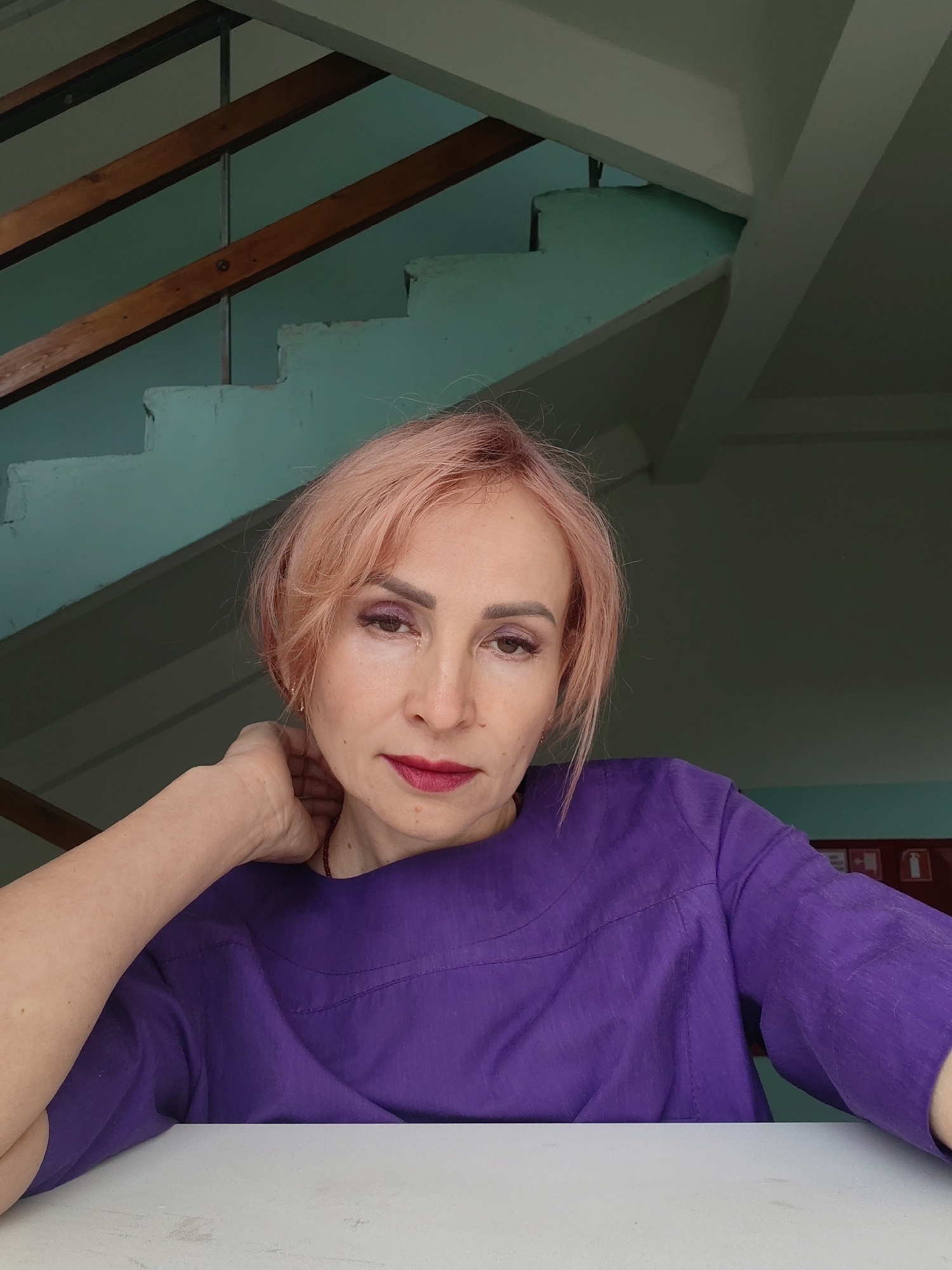 A fair-skinned woman with pink hair and dark lipstick is looking at the camera. She is wearing a purple shirt and her right arm is bent with her hand behind her neck. She is sitting at a white table. In the background, a light blue wall with a staircase and wooden railings is visible. The image appears to have been taken indoors, possibly in an apartment building or a public space, but there is no specific information to determine the location within Chaikovsky, Russia.