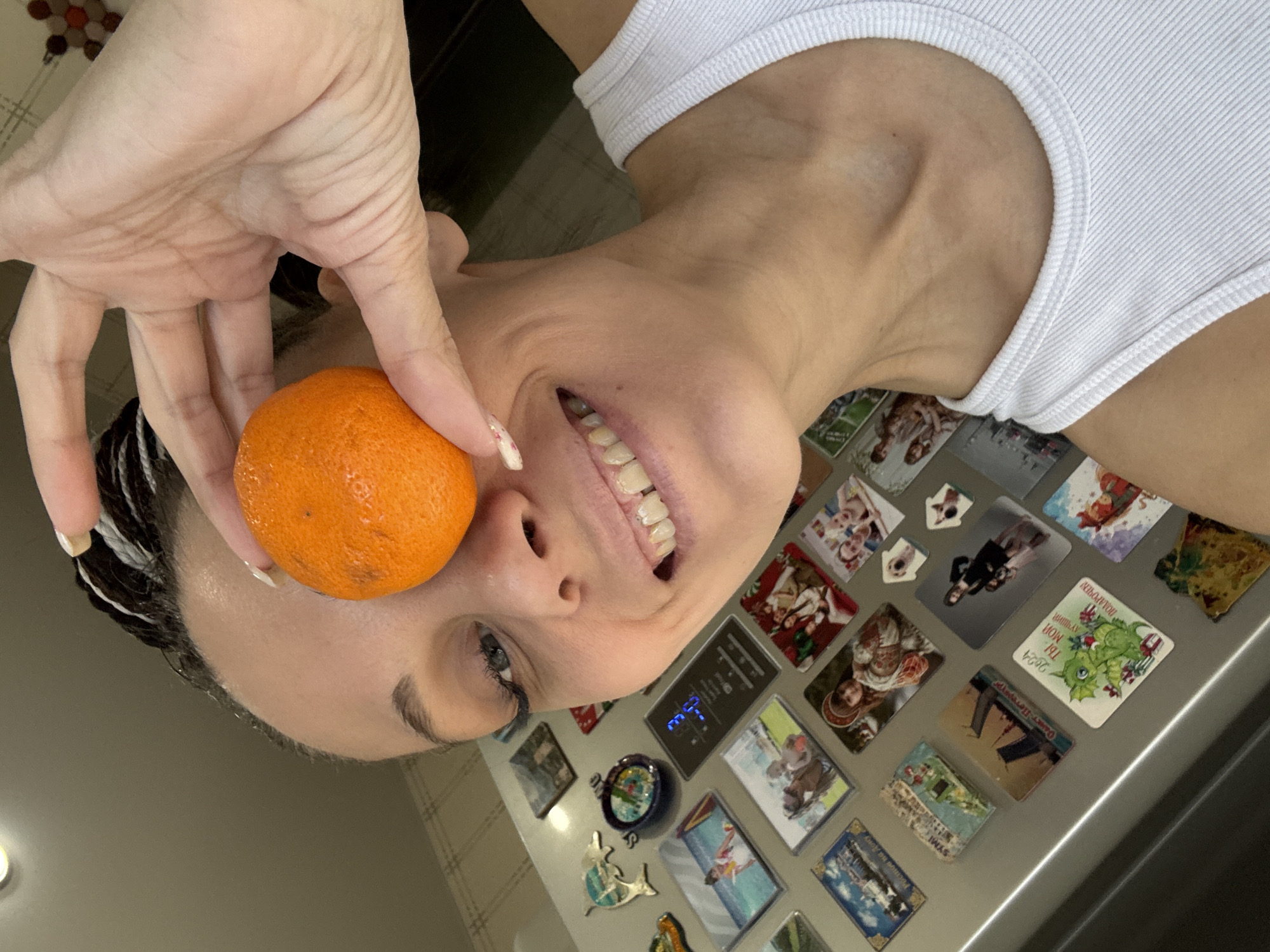 A woman holds an orange in front of her left eye while standing in front of a refrigerator covered in magnets. She wears a white tank top and smiles. Her black hair has white streaks. The magnets on the refrigerator display various images. The location is assumed to be Rusanovo, Russia, based on context not present in the image.