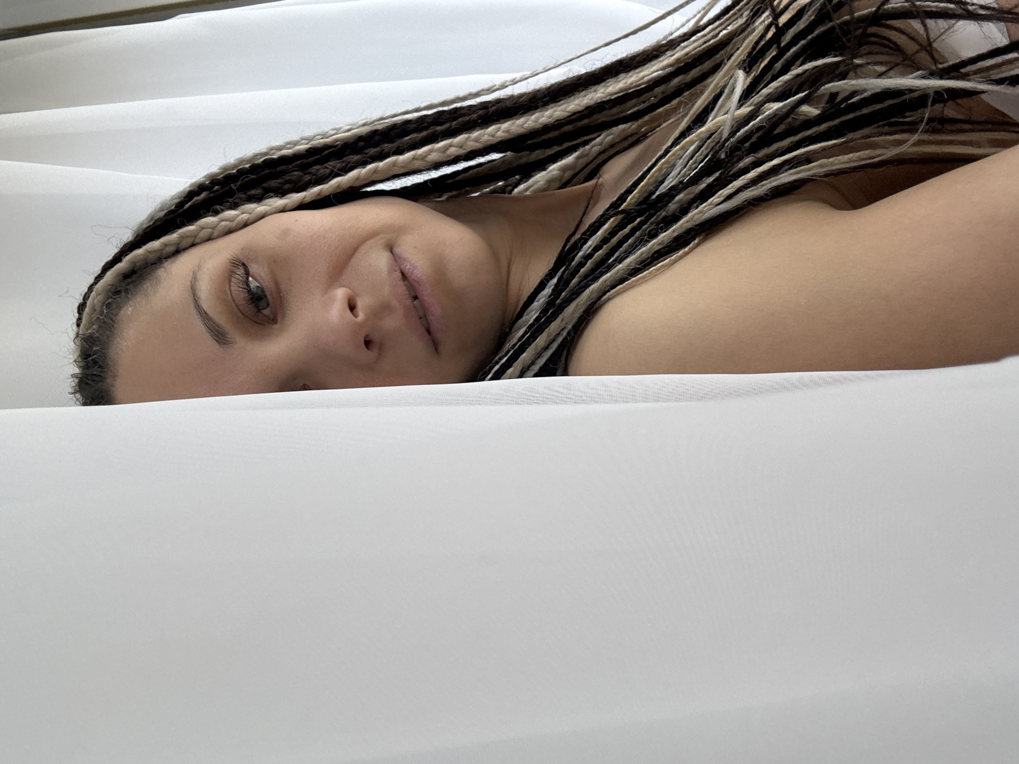 A woman with braided hair is lying down on a white surface. Her head is tilted towards the camera, and she is smiling slightly. Her skin tone appears light. The environment around her appears to be a white, soft material. The city and location are unknown.