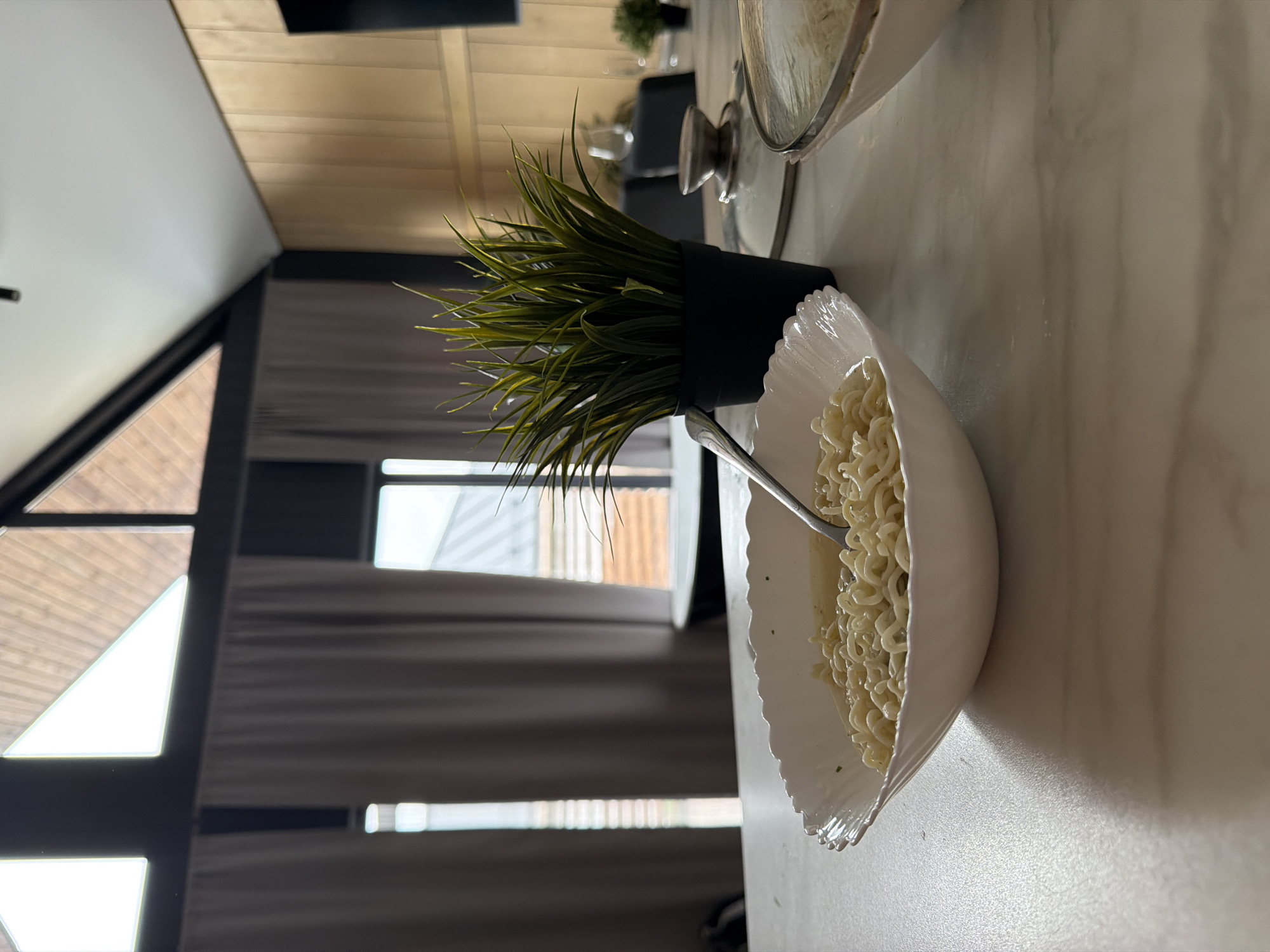 A close-up, eye-level shot captures a bowl of ramen noodles with a fork resting in it, placed on a marble-patterned countertop. To the right, a potted green plant adds a touch of life to the scene. In the background, a modern interior features large triangular windows with grey curtains, a wooden-paneled wall, and other dining elements like glasses and a lid from another pot. The overall impression is one of a casual meal in a contemporary living space. The location is identified as Semiluki, Russia.