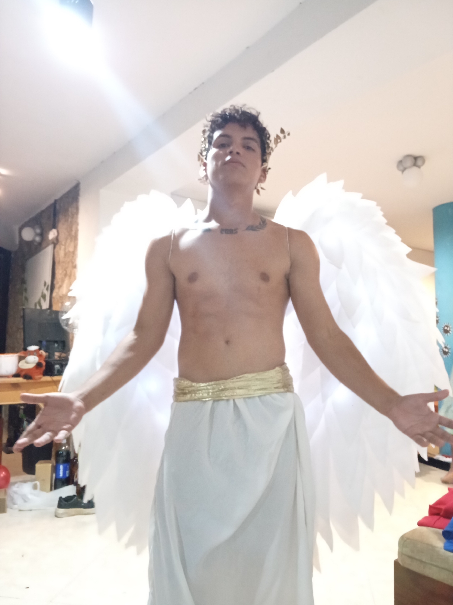 A young man stands indoors, dressed in an angel costume. He has bare chest and is wearing a white toga-like garment with a gold sash. Large white wings are attached to his back, and he has a gold leaf crown on his head. His arms are extended to the sides. The location is Playa El Agua, Venezuela. Background details include furniture, a television, and a blue-painted wall.