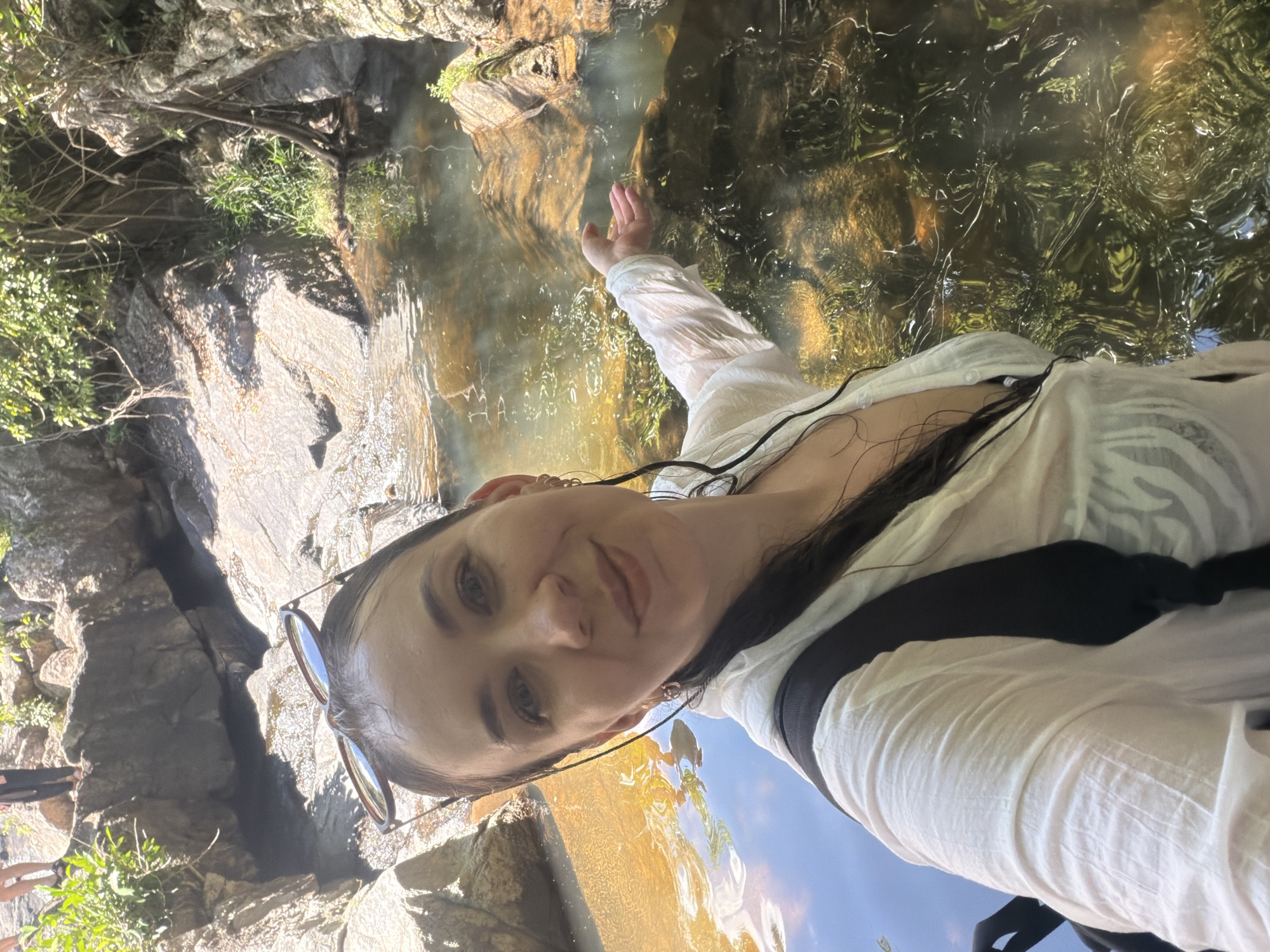 In a selfie taken at a natural waterfall in Hoa Thang, Vietnam, a woman with dark, wet hair and sunglasses perched on her head is centered in the frame. She is wearing a sheer white shirt, partially unbuttoned, revealing a dark bra underneath. A black backpack is visible over her shoulders. Her right arm is extended towards the water, with her palm open, as if testing its temperature or interacting with it. The environment features a shallow, clear body of water with a rocky bed, surrounded by large, moss-covered rocks and lush green foliage. Sunlight filters through the trees, creating dappled patterns on the water's surface. In the background, two individuals are standing on rocks at the top of the waterfall, partially obscured by vegetation.