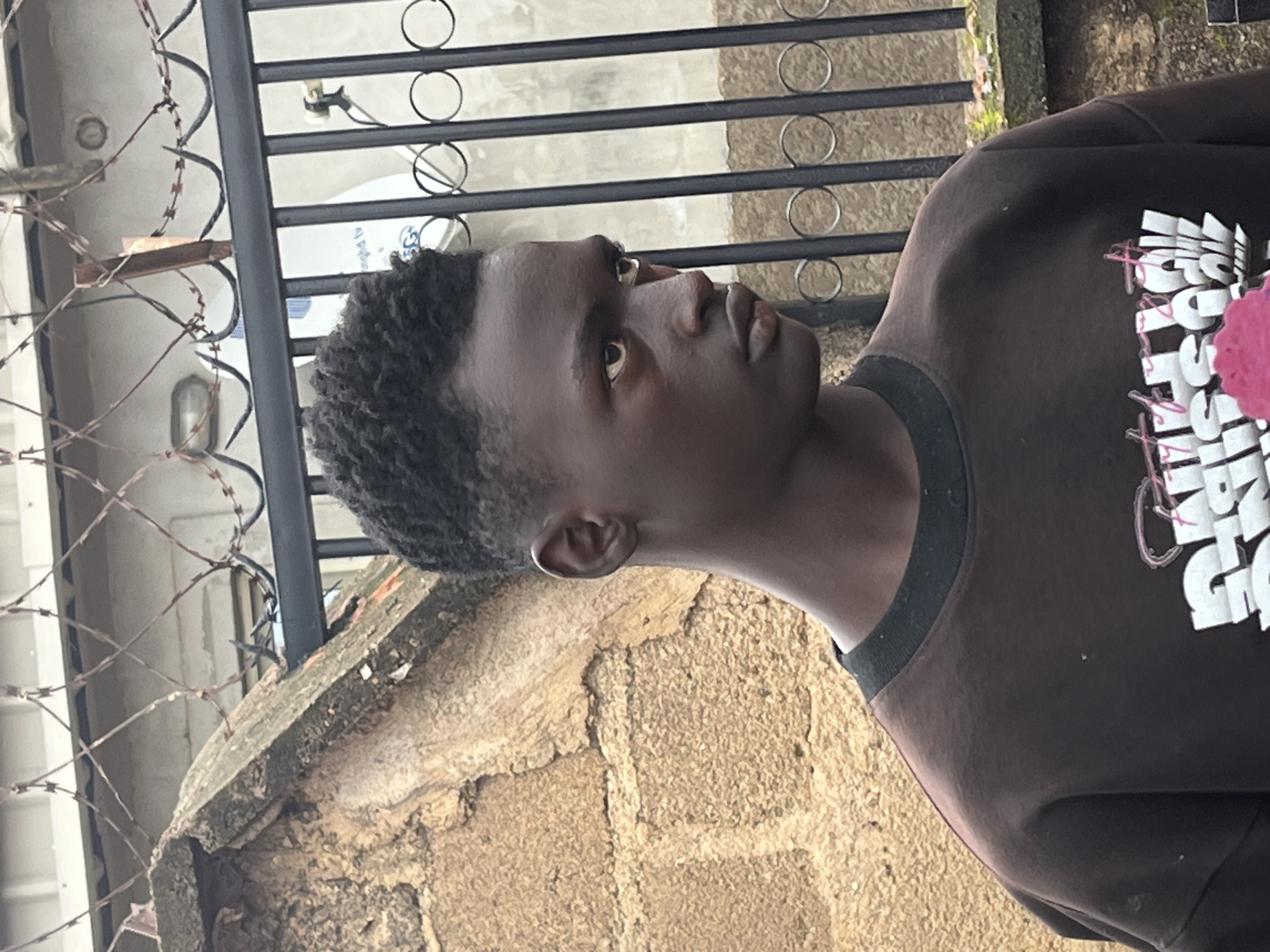 A medium shot captures a young Black male from the chest up, facing right, with his head turned slightly towards the camera. He has dark skin, short, curly dark hair styled upwards, and is wearing a dark brown crewneck sweatshirt with white and pink text and graphics on the front. He is standing outdoors, with a rough, textured tan brick wall behind him on the left, and a black metal fence with circular decorative elements and razor wire on top, partially obscuring a light-colored background with a light fixture and what appears to be a white object with blue writing. The scene is lit by diffused daylight.
