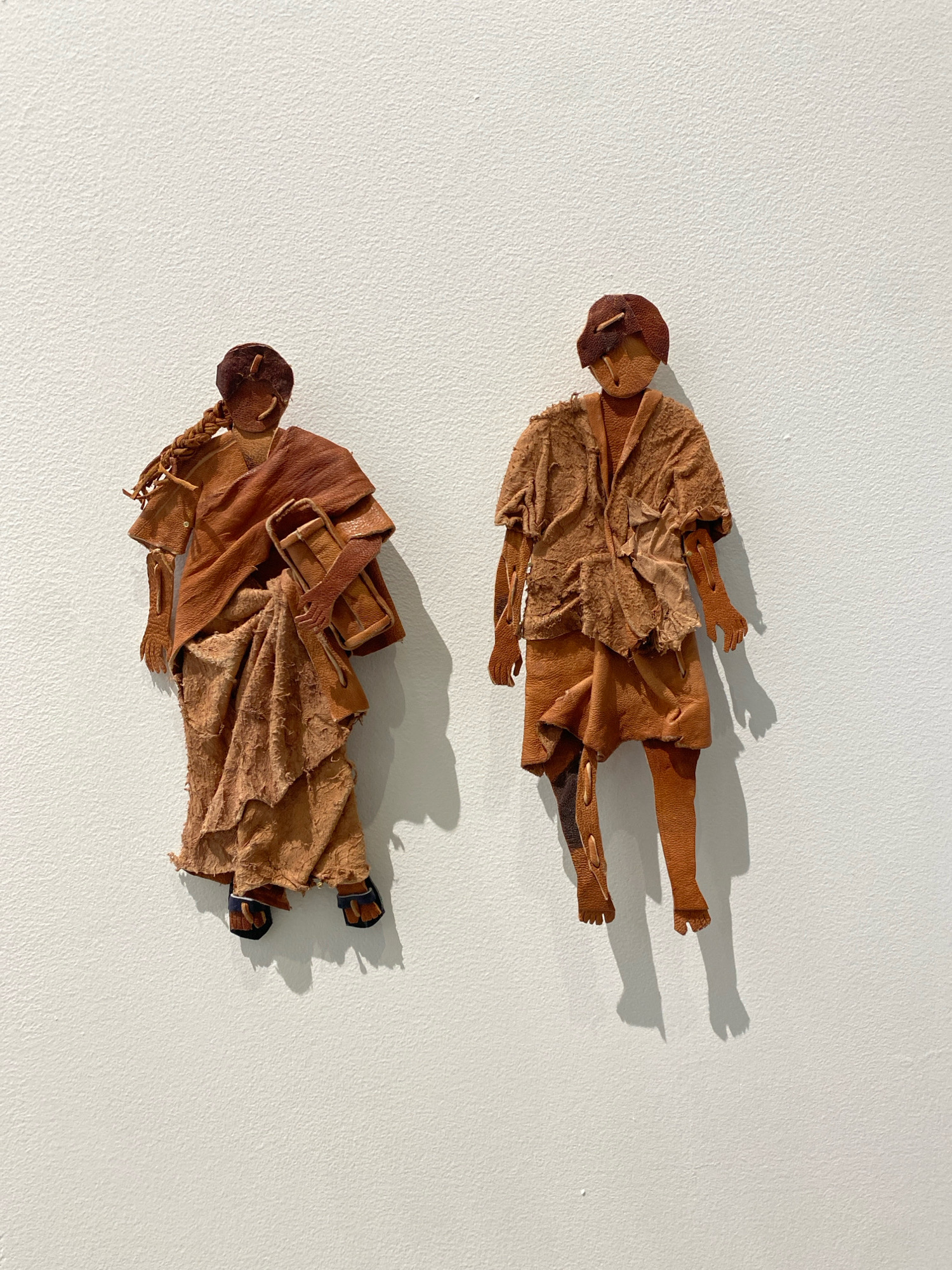 Two abstract, humanoid figures, constructed from what appears to be distressed leather or fabric in shades of brown and tan, are mounted on a white textured wall. The figures are positioned side-by-side. The figure on the left has its hair styled in a braid and is holding a cylindrical object in its right arm. It is wearing a long, layered garment and sandals. The figure on the right wears a shorter, similarly layered outfit, revealing its legs. Both figures have simple, abstract facial features. Shadows are cast by the figures on the wall, indicating light from the right. This image was captured in an indoor setting, likely an art exhibition, and is not located in the city of Khimki, Russia.