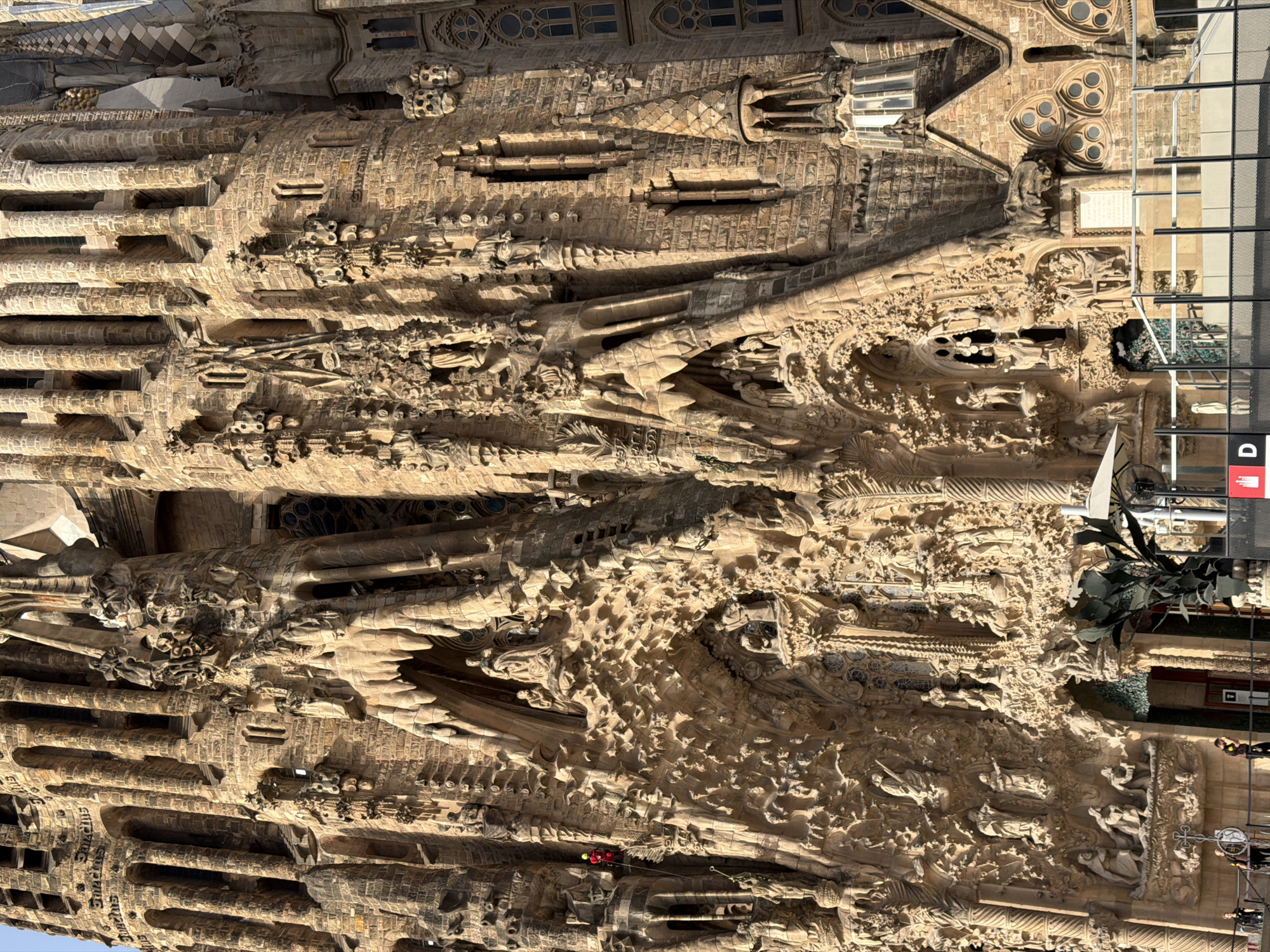 The image shows a detailed view of the Sagrada Familia basilica in Barcelona, Spain. The basilica's facade is elaborately decorated with intricate sculptures, depicting religious scenes and figures. The stone architecture is textured and weathered, with sunlight casting shadows that highlight the depth and detail of the carvings. In the middle left of the image, a person in a red and yellow outfit is visible, appearing to be rappelling down the side of the building, possibly for maintenance or cleaning. Below this, on the ground level, several people are standing near a metal fence. The lower part of the image also shows part of the entrance area, with some protective coverings and signage. The sky is not visible, as the focus is entirely on the facade of the basilica.