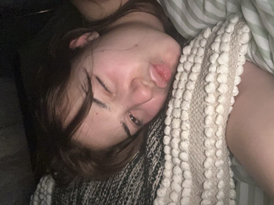 A young woman is lying down, close to the camera. Her face is angled towards the viewer, with one eye partially closed. She has brown hair and fair skin, with a slightly pouty expression. Her head rests on a white and tan patterned blanket, with a green and white striped fabric visible behind her. Part of her arm is visible next to the blanket. The background to the left is dark.