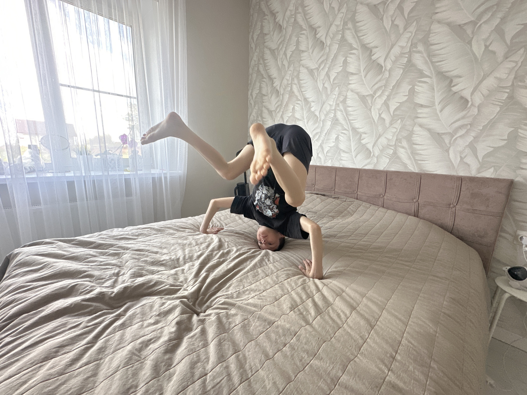 A boy is doing a headstand on a bed with a beige comforter. He wears a black t-shirt and dark shorts. His head is touching the bed, and his hands are providing support. One leg is raised straight into the air, while the other is bent. The bed has a headboard made of a beige, textured material. The background includes a window with sheer white curtains, revealing a view of what appears to be a residential area. The wallpaper features a repeating pattern of leaf-like shapes in a light neutral tone. A small white side table with objects sits on the right side of the image. It's likely this image was taken in Прикамский, Russia.