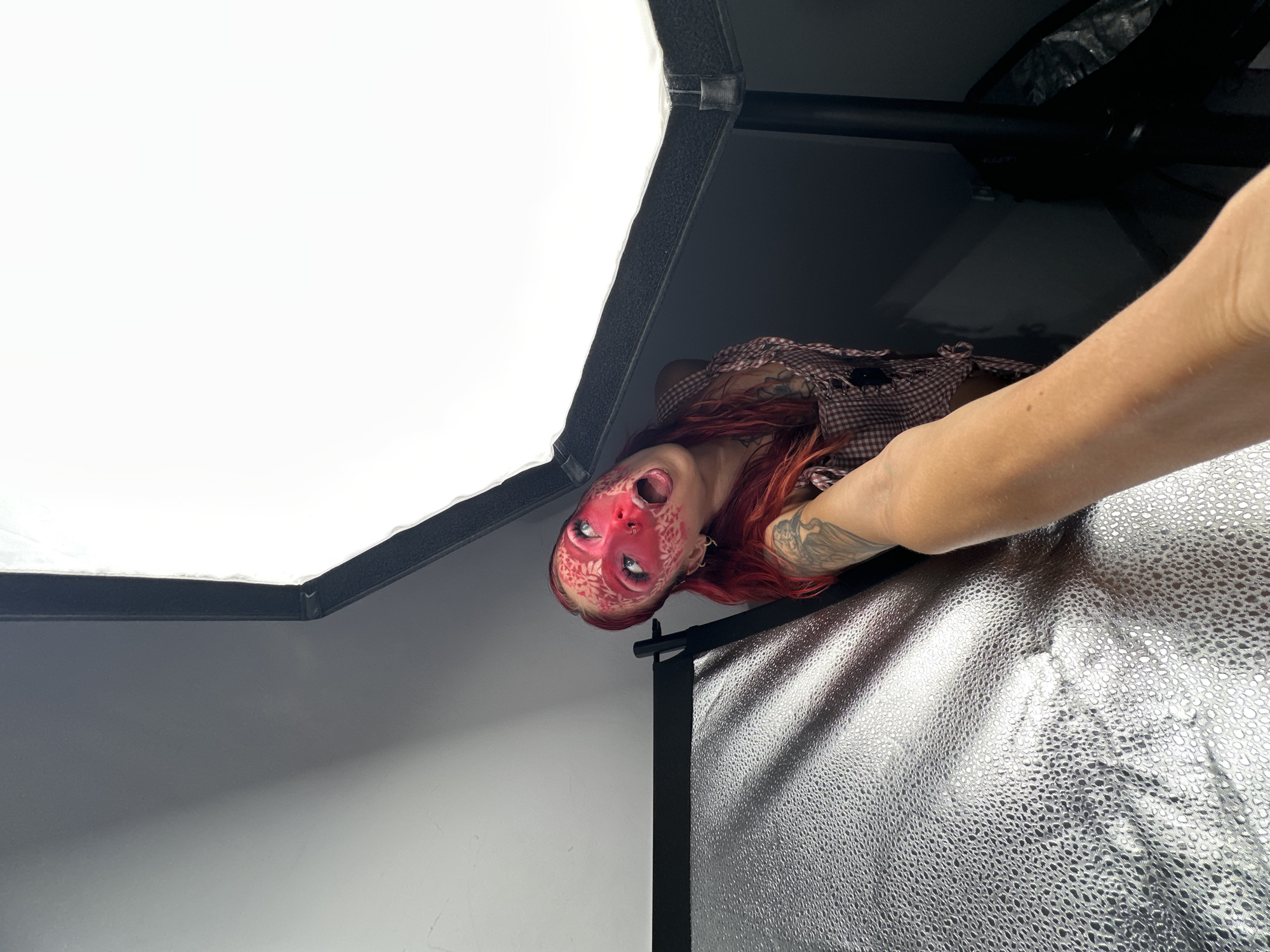 A woman with red hair and intricate red and white face paint resembling scales is posing in what appears to be a photography studio. She is looking up and to her left with her mouth open. She is wearing a red and white checkered vest. A hand is reaching into the frame from the lower left corner, holding the camera. A large softbox light is visible in the upper right portion of the frame. A silver reflector is also visible on the left side of the frame. The background is dark and out of focus. There are no identifiable landmarks or features that would place the scene in North Kuta, Indonesia.