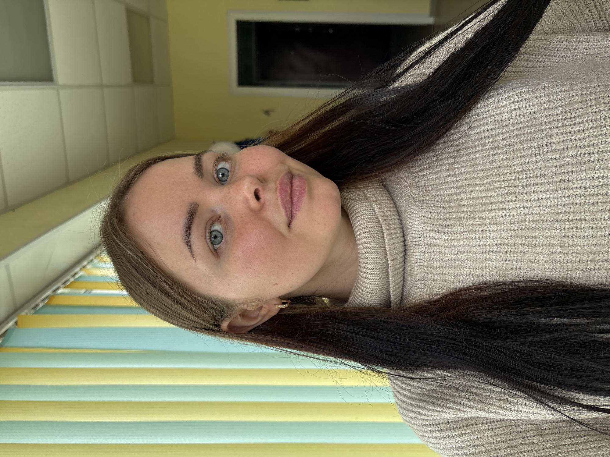 An indoor portrait shows a young woman looking directly at the camera, with her head slightly tilted. She has fair skin with visible freckles, light-colored eyes (blue or grey), and a neutral expression that might suggest a faint smile or surprise. Her long, straight hair is predominantly dark, with lighter brown or blonde strands framing her face. She wears a light beige or oatmeal-colored knitted turtleneck sweater. A small, golden hoop earring with several tiny stones is visible in her right ear.
The background on the left side features vertical blinds displaying alternating horizontal stripes of light blue and yellow-orange. On the right side, a plain light yellow wall is visible, above which a dark rectangular object or opening, possibly a framed picture or vent, can be seen with some light reflections. Further up, there is a light-colored wall section, potentially tiled or paneled. The setting is a well-lit indoor environment. No text is visible in the image. The activity is a self-portrait.