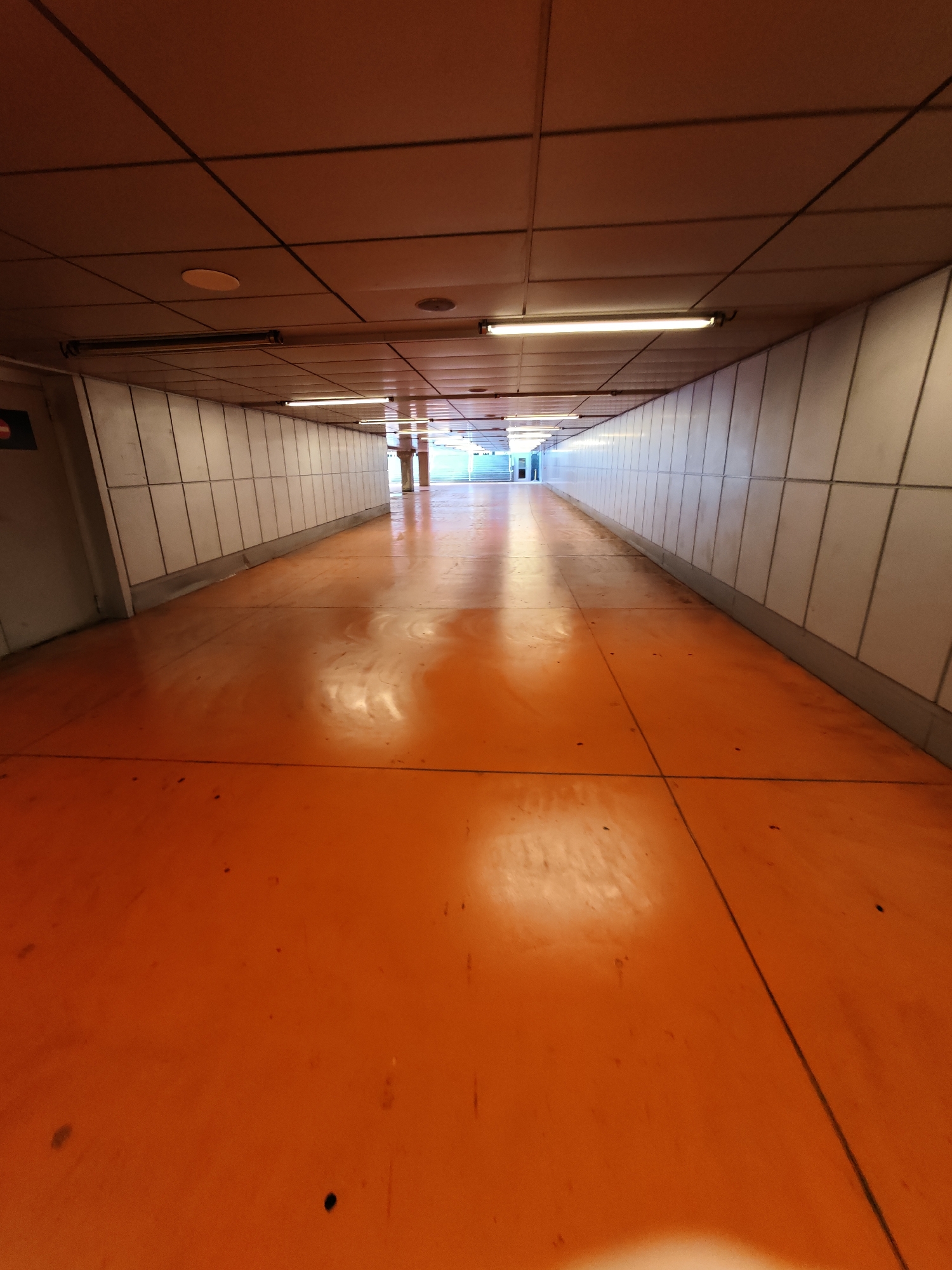 The image shows an empty, brightly lit pedestrian tunnel with an orange floor and white tiled walls. The tunnel is lined with fluorescent lights on the ceiling. The perspective suggests the image was taken from a low angle looking down the length of the tunnel. The environment appears to be a modern, possibly public, transit area. There are no people or any discernible actions or interactions. The scene is clean and well-maintained, with reflections visible on the polished orange floor.