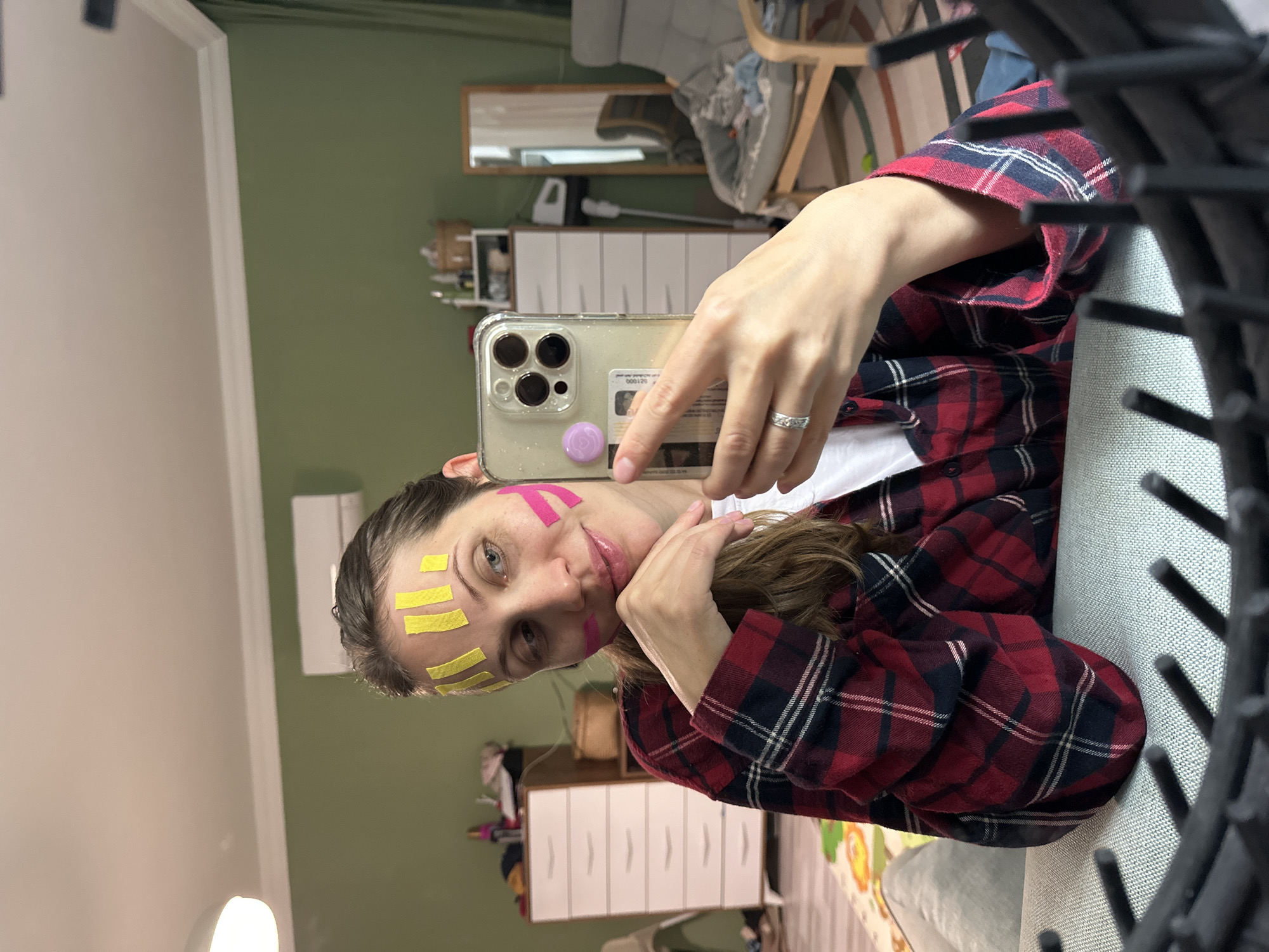 A woman is indoors taking a selfie in front of a light green wall. She is wearing a red and black flannel shirt and has yellow and pink tape on her forehead and cheek. She is holding a phone with a clear case that has a purple circle on the back, and she is wearing a ring on her finger. A black object with vertical bars is in the foreground, partially obstructing the view. In the background, there is a white dresser, a mirror, and a chair with a gray cushion. The location of the scene is not explicitly available. The city of Fethiye, Turkey is not explicitly known from the content of the image.