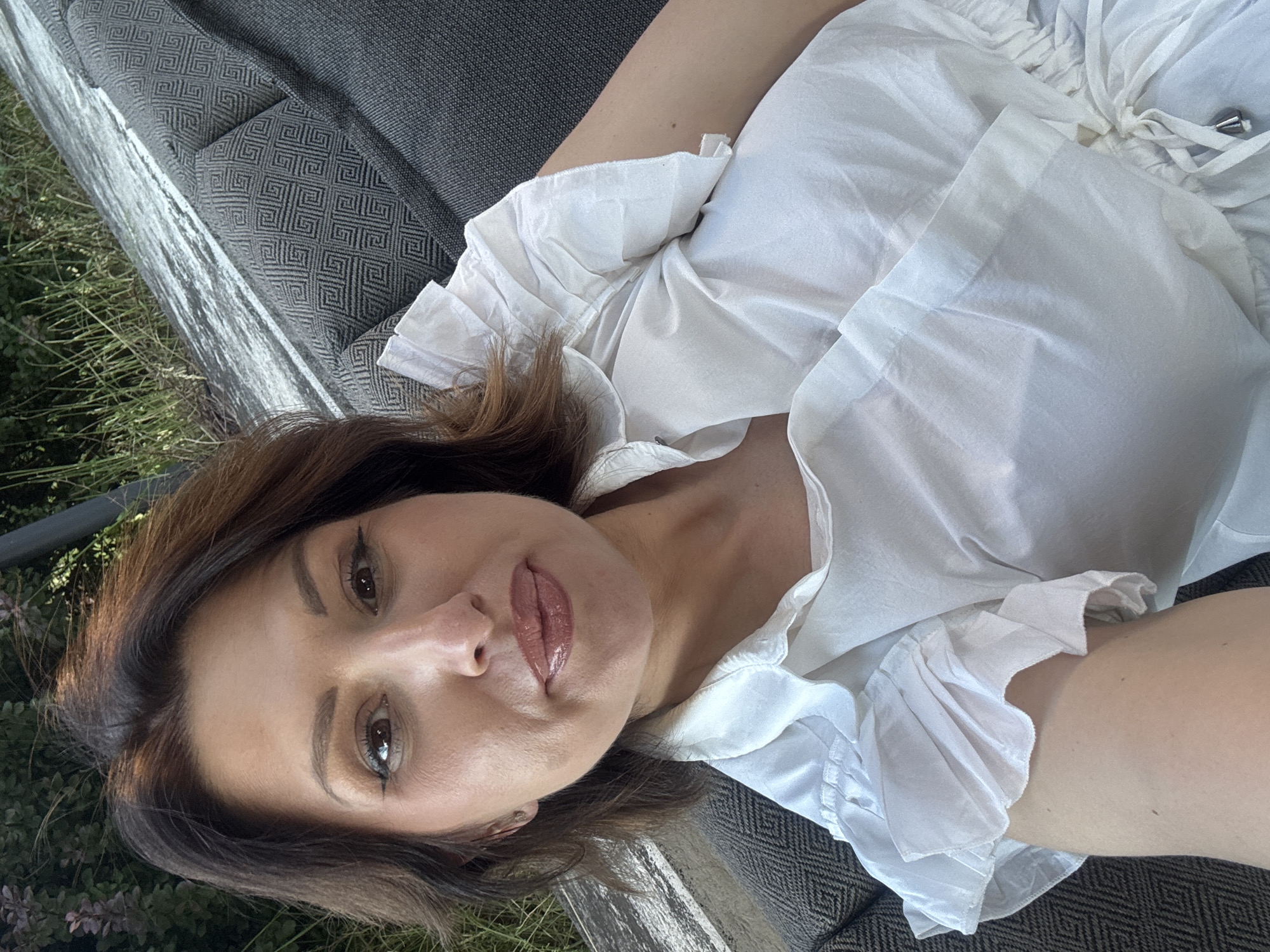 A woman with brown hair and fair skin, wearing a white ruffled blouse, takes a selfie while sitting on an outdoor couch. The couch has a dark gray fabric with a geometric pattern. Behind the couch, there is a textured, weathered surface and some greenery. The overall scene suggests an outdoor setting, possibly a garden or patio, in Sofia, Bulgaria.