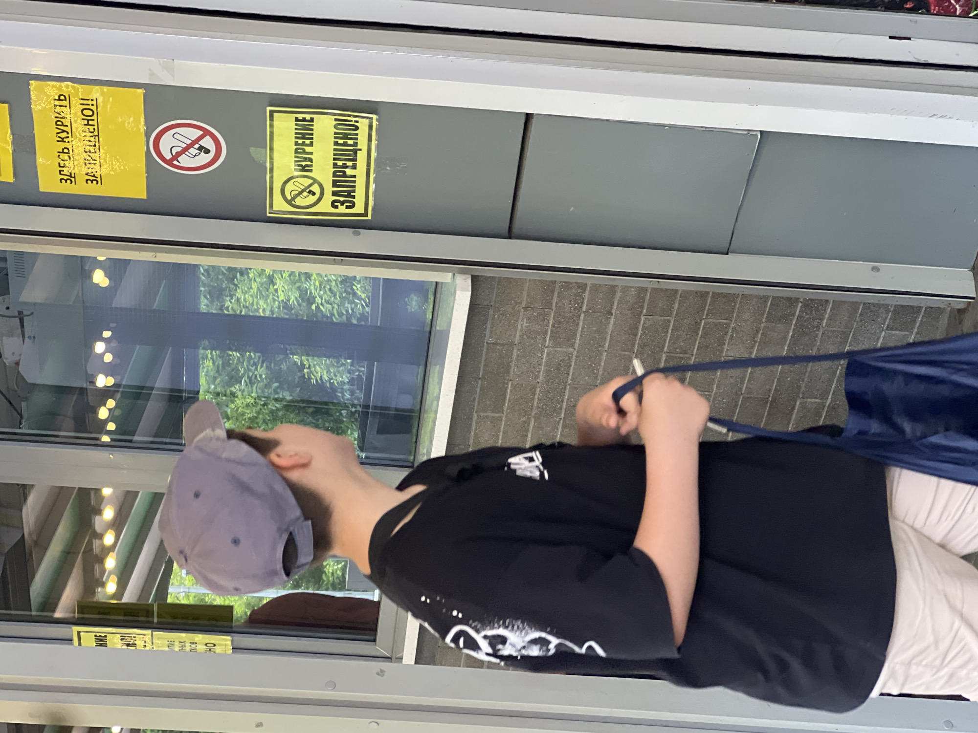 In Sochi, Russia, a young person wearing a grey baseball cap and a black t-shirt with white graphics is standing next to a glass window. They are holding a blue bag in their right hand and a cigarette in their left hand. The person is looking towards the window, which has several yellow signs posted on a grey surface to the right. One sign has a red circle with a diagonal line over a cigarette, indicating no smoking. Another yellow sign reads "КУРЕНИЕ ЗАПРЕЩЕНО!" which translates to "SMOKING IS PROHIBITED!". The environment appears to be outdoors, possibly near a kiosk or service window, with greenery visible through the glass.