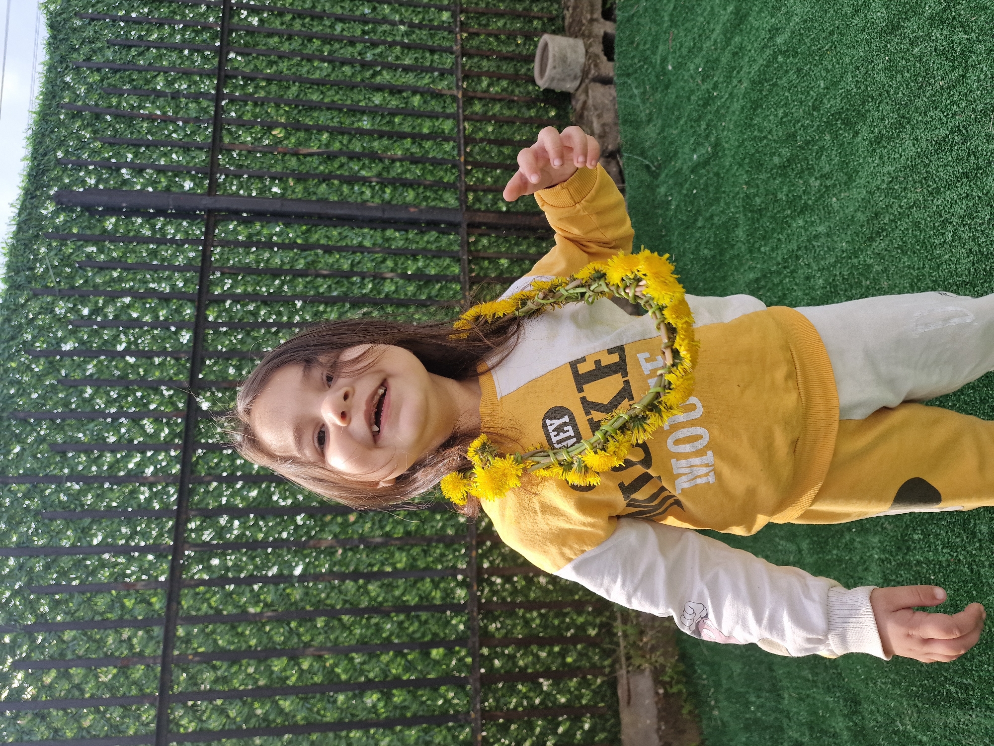 A young girl stands in a garden. She wears a yellow and white sweater with the word "MOUSE" printed on it and a garland made of yellow dandelions. Behind her is a fence covered with artificial green foliage. The ground is covered in artificial grass.