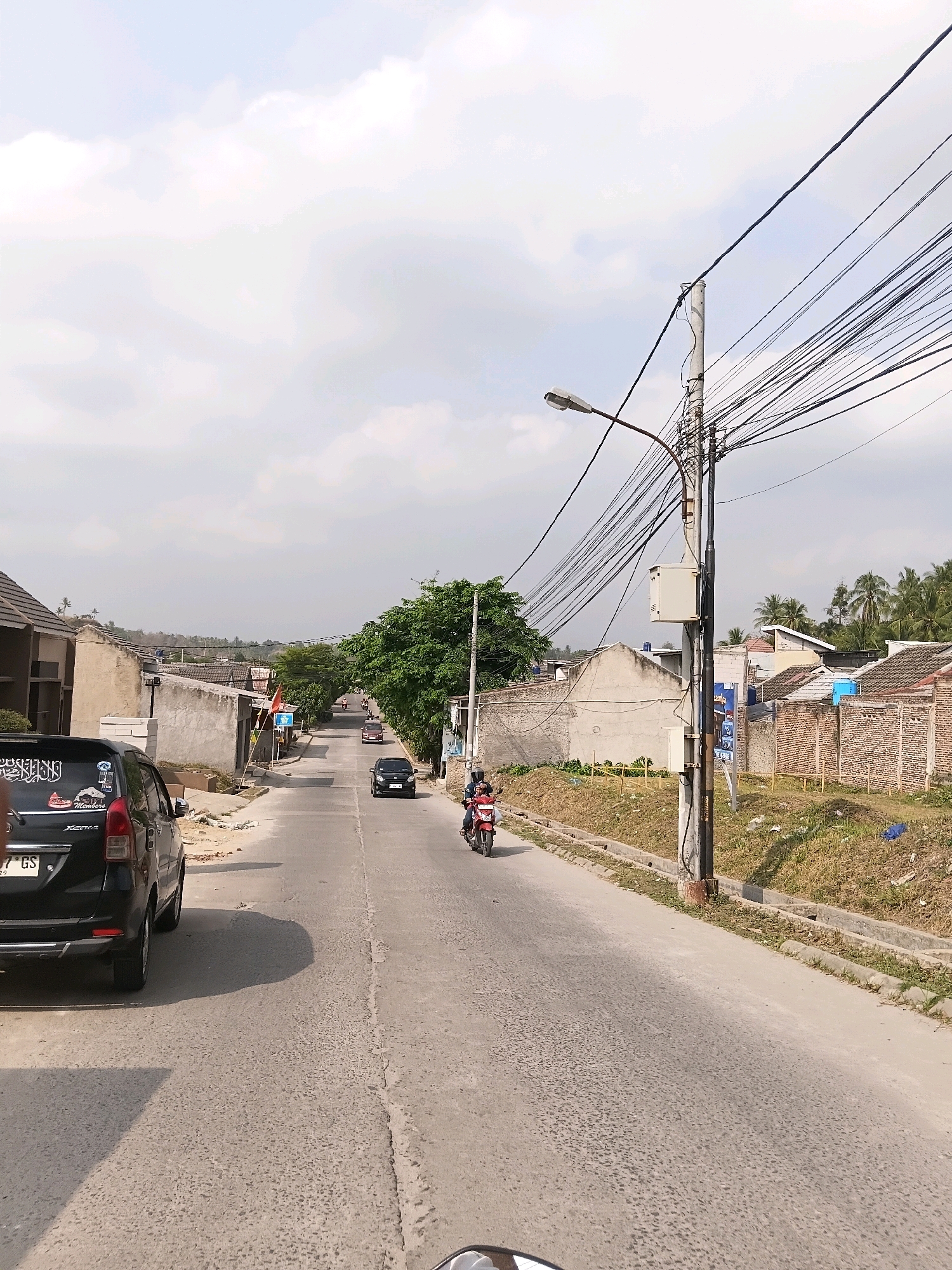 The image shows a street scene in the city of Cilegon, Indonesia. A black car is parked on the left side of the road, and a motorcycle with two riders is visible further down the road, moving away from the viewer. The street is paved, and there are houses and buildings on both sides of the road. A tall utility pole with numerous electrical wires and a street light is prominently featured on the right side of the road. The sky is overcast with clouds. The vegetation includes trees and dry grass.