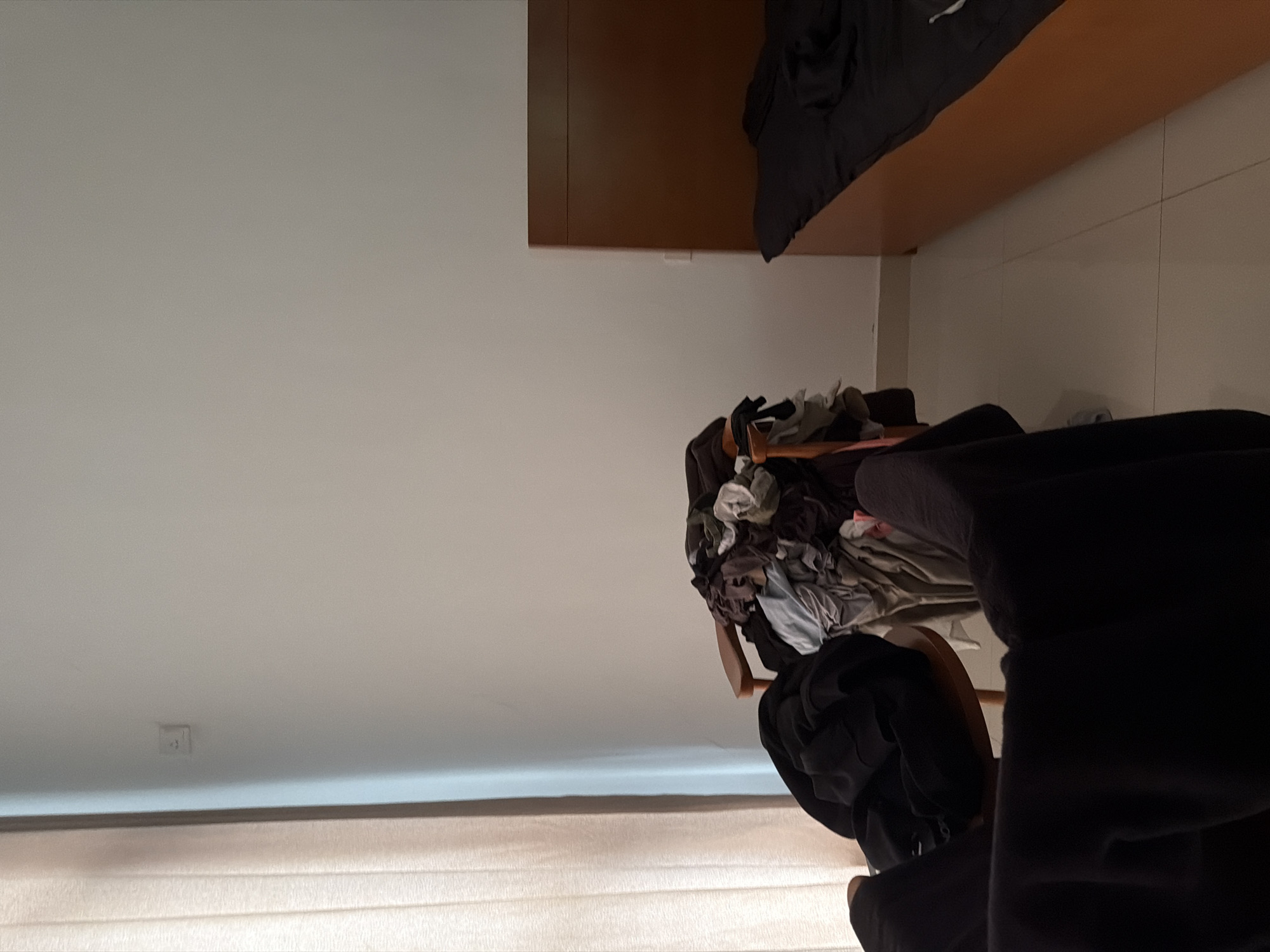 The image shows a room interior. A pile of clothes sits on a chair. Above the chair is a brown shelf or cabinet. The background features a beige curtain and a light-colored wall. The location is Jiangning District, China cannot be determined from the image.