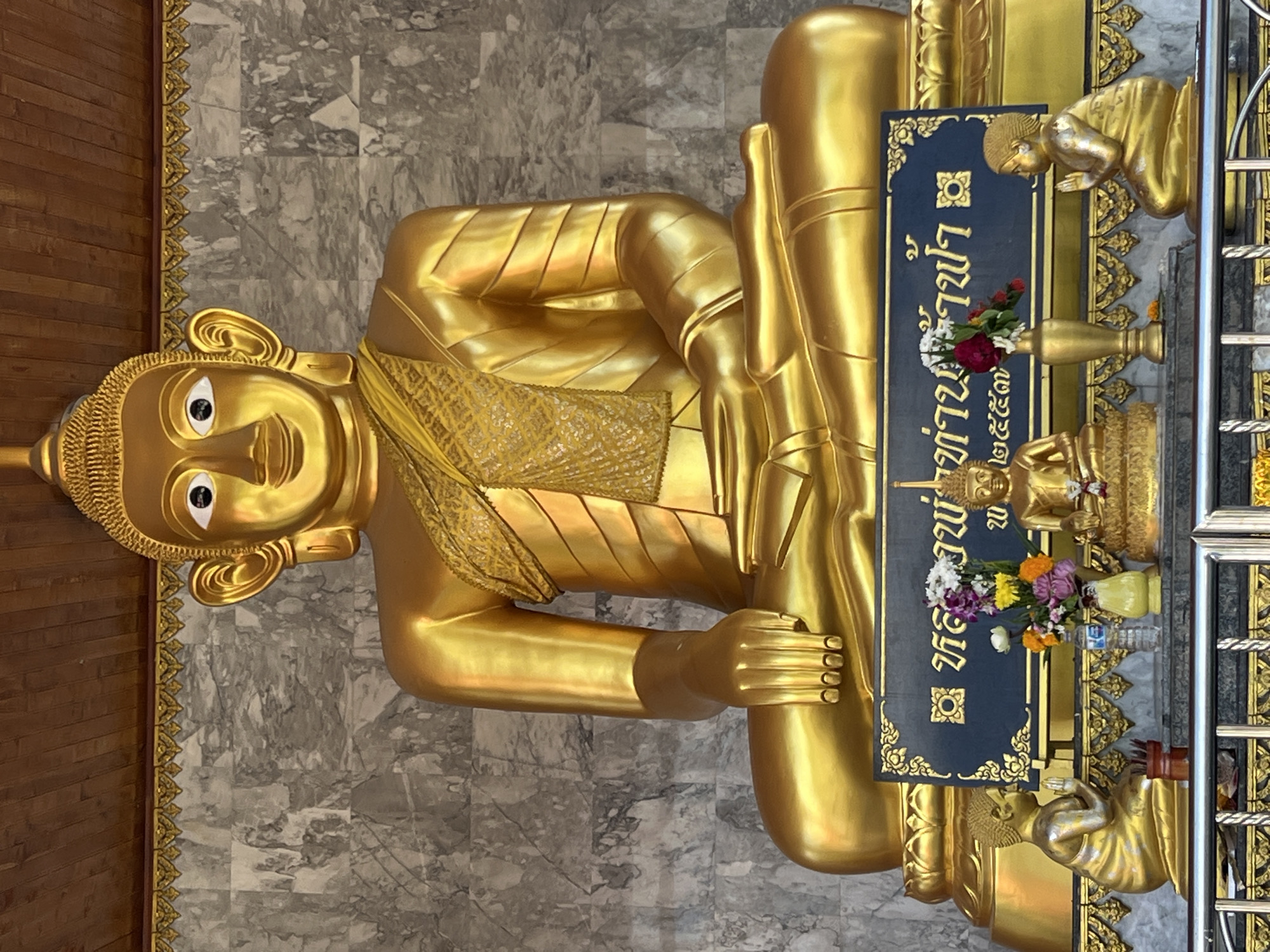 The image depicts a large, golden Buddha statue seated in a meditative posture, with its hands resting on its lap. The statue is centrally placed in a shrine, with a marble-patterned wall behind it and a wooden ceiling above. Flanking the main statue are two smaller golden figures of devotees in prayer. In front of the main Buddha, a dark blue plaque with Thai inscriptions and gold trim is displayed, adorned with a floral arrangement. Below the plaque, a smaller golden Buddha statue is visible, also surrounded by offerings. The scene appears to be located in Thep Krasatti, Thailand, suggesting it is a religious or cultural site. The overall atmosphere is one of reverence and tranquility.