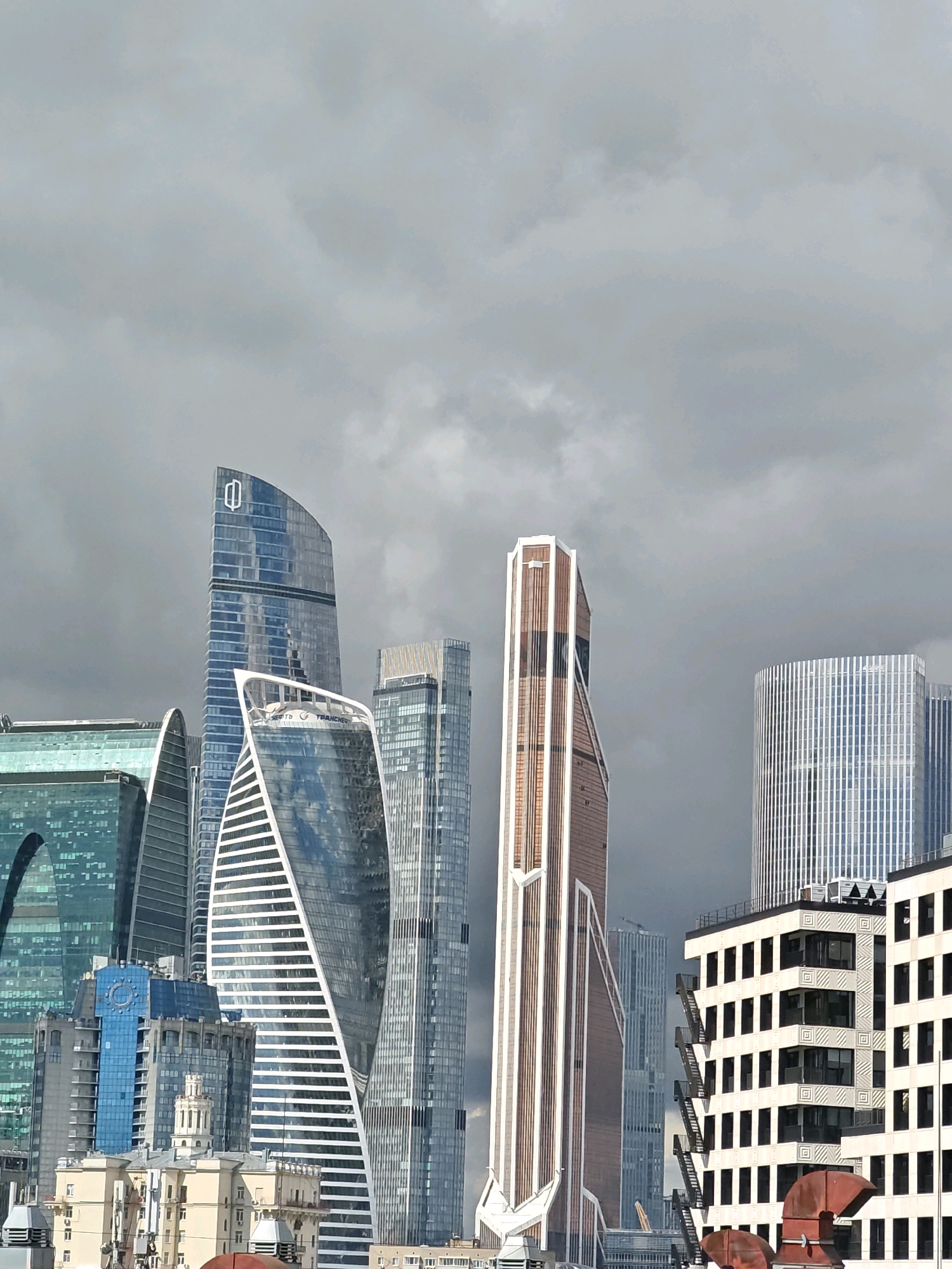 The image shows a collection of modern skyscrapers in the Moscow International Business Center, also known as Moscow City, Russia. The sky is overcast and filled with dark clouds. The buildings are diverse in their design and materials, with glass facades reflecting the sky and surrounding structures. Some prominent buildings include the towers with twisted shapes, a building with a copper-colored facade, and a cylindrical tower with a grid-like exterior. In the foreground, there are some lower-rise buildings, appearing to be older architecture, with visible rooftops and details. The overall scene depicts a dense urban environment with a focus on contemporary architectural styles. There are no people visible in the image.