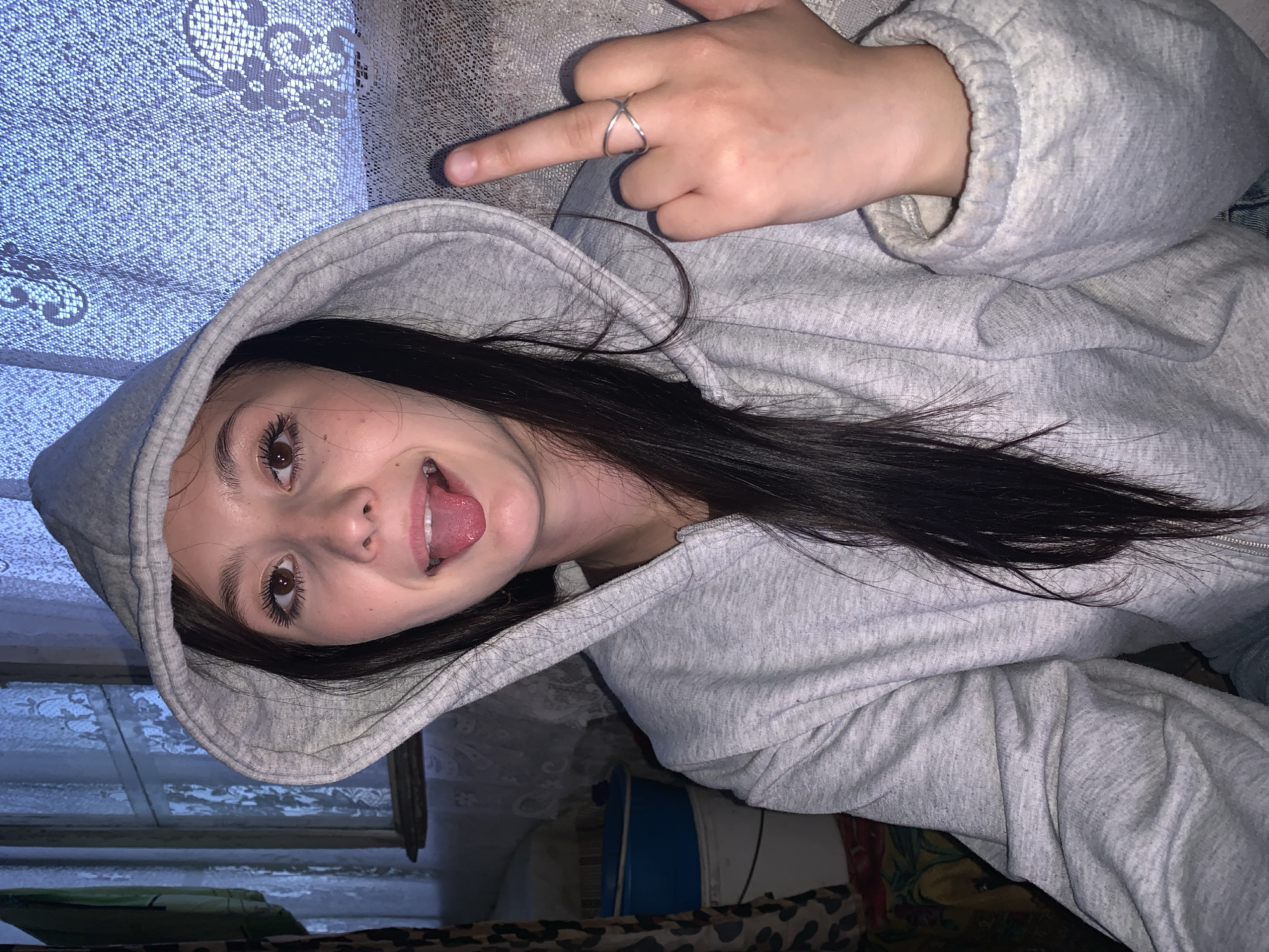 A young person, likely female, is depicted in a close-up, tilted portrait, possibly a selfie, with a playful expression. Her head is angled, and she is sticking out her tongue. She has dark hair, brown eyes with long eyelashes, and is wearing a light grey hooded sweatshirt with the hood partially up.
Her left hand is visible in the upper right portion of the image, with her index finger extended in a casual gesture. A silver criss-cross ring is worn on this finger. The sleeve of her grey hoodie is also visible.
The setting appears to be indoors, characterized by varied textures and patterns in the background. On the left, a light blue or grey fabric with a distinct white lace or floral pattern is present, resembling a curtain or bed linen. The area behind her body and to the right features a mix of textures, including what looks like part of a blue and white container and a brown and black animal-print fabric.
The lighting is somewhat dim, suggesting an indoor environment or evening time. The overall mood conveyed by the subject's expression is casual and cheeky. There is no visible text in the image. The location context provided is Fokino, Russia.