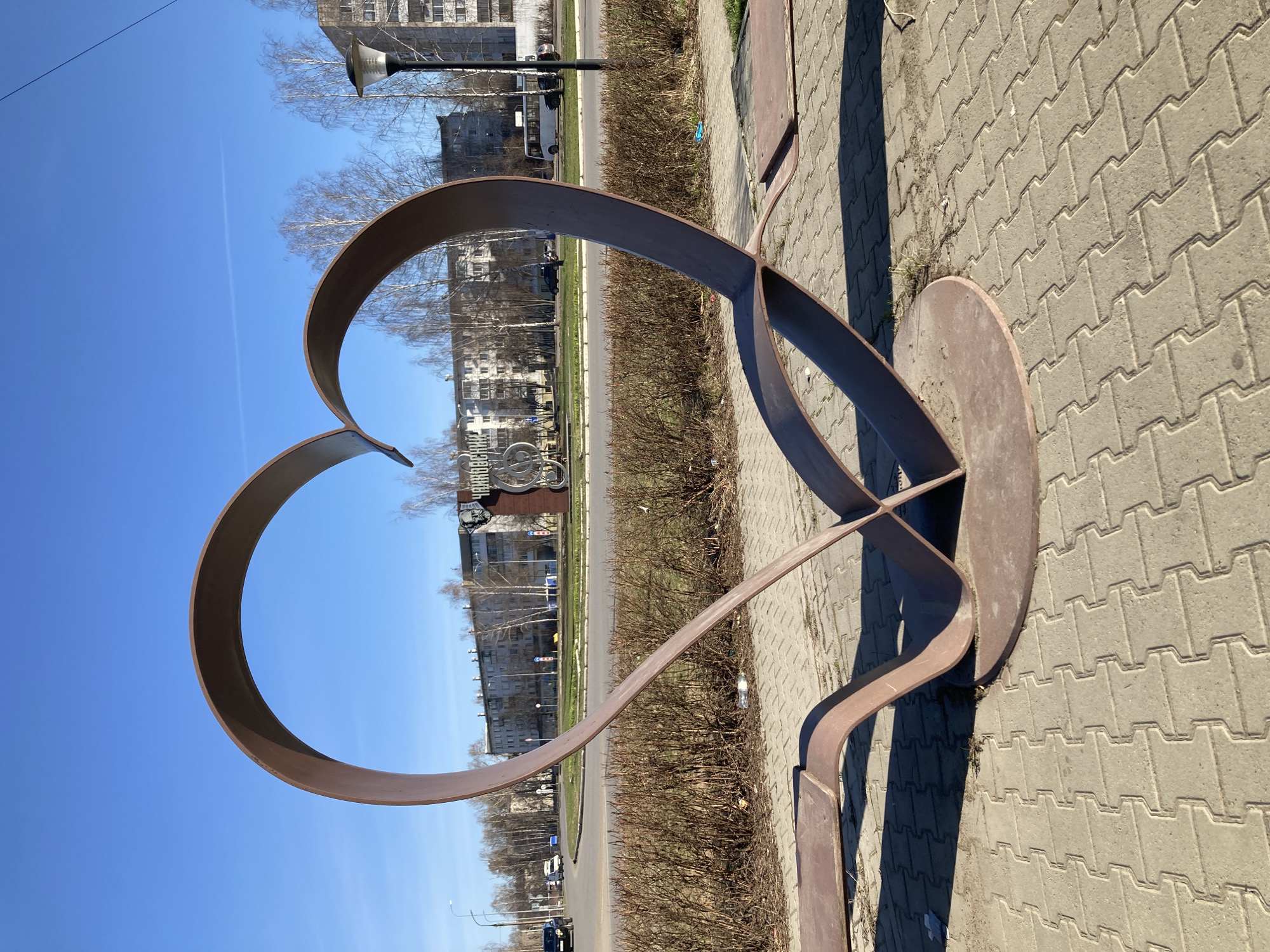 The image shows a large metal heart sculpture located on a paved walkway next to a street in Chaikovsky, Russia. Behind the sculpture, there is a line of bushes, a road with vehicles, and buildings in the background. The sky is clear and blue. The sculpture is made of a wide metal band shaped into a heart, supported by a base and additional metal structures. The walkway is made of gray paving stones.