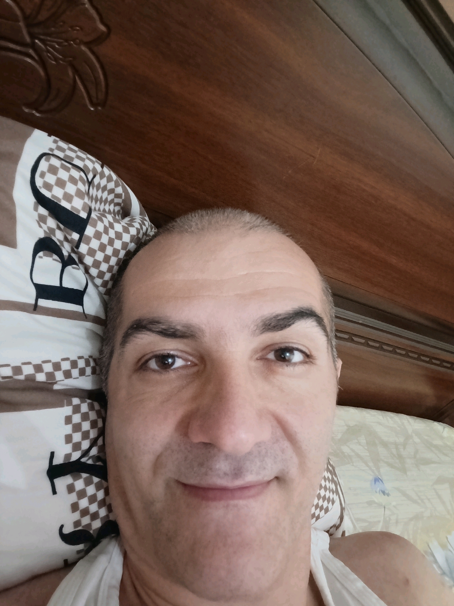 The image is a close-up selfie of a man lying in bed. He has short, dark hair, dark eyebrows, and brown eyes. He is smiling faintly. The man is wearing a white tank top. The background shows a pillow with a checkered pattern and a black mustache design, and a wooden headboard of a bed. The scene appears to be indoors, in a bedroom. There are no discernible features that would indicate the location is in Grozny, Russia.