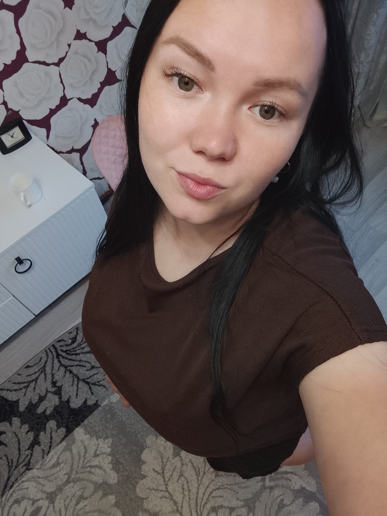 A woman poses for a close-up indoor selfie, looking directly at the camera with a subtle smile, in Kuyeda, Russia. She has long, dark hair and light brown or hazel eyes. She is wearing a brown, ribbed, short-sleeved t-shirt.
The background features a white nightstand with a black ring-shaped drawer pull. On the nightstand, a small black-framed analog clock and a white mug are visible. The wall behind is adorned with a distinctive wallpaper pattern of large white roses on a dark purple background. A pink-upholstered chair is partially visible to the right of the nightstand. The lower portion of the image shows a patterned rug on the floor, featuring grey, black, and light grey floral or leafy motifs. The lighting is soft and even, typical of an indoor setting. No text is visible in the image.