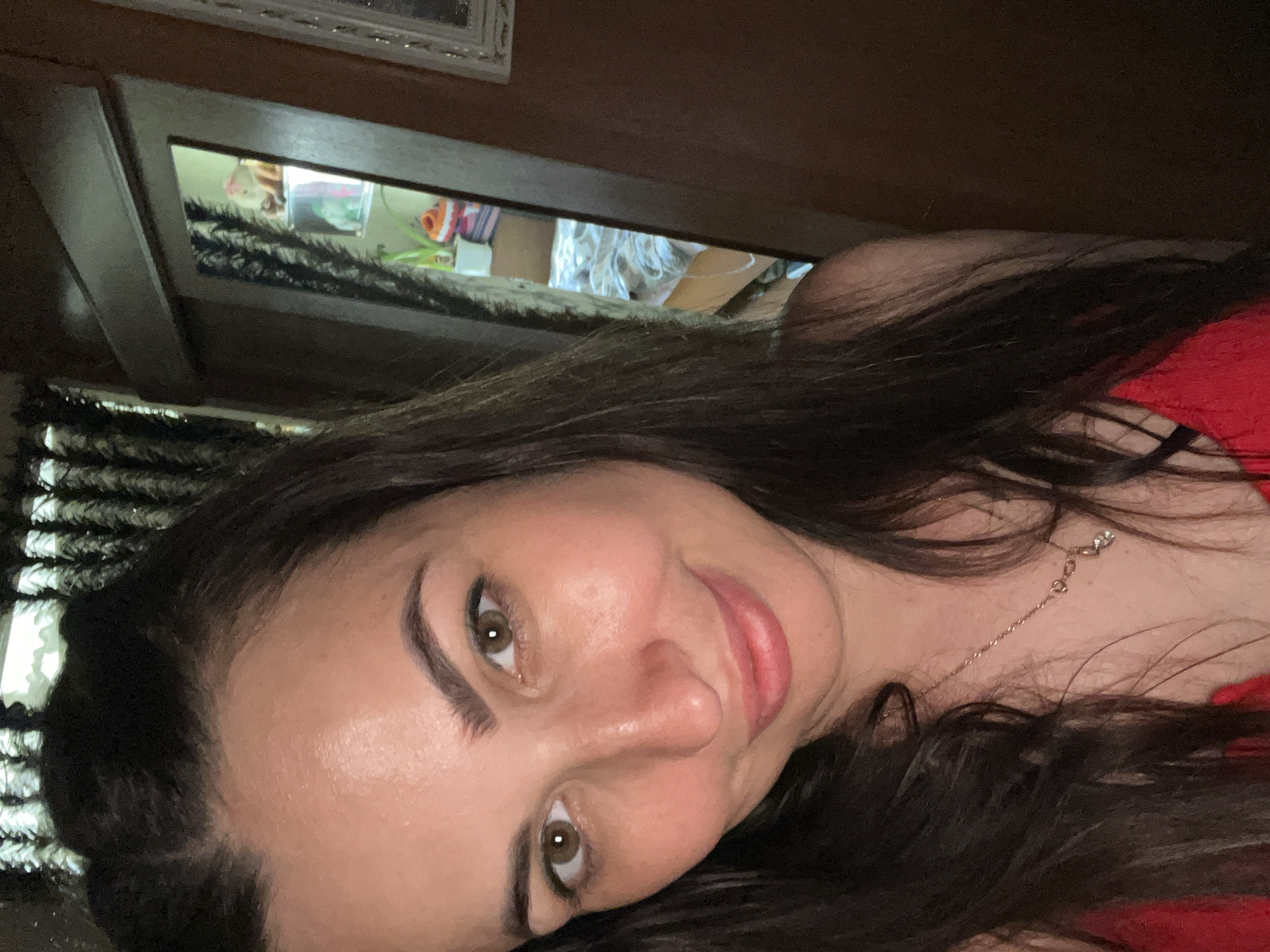 A woman is taking a selfie in a room. She has long, dark brown hair and brown eyes. She is smiling and wearing a red top and a necklace. The background includes a mirror that reflects a bed and some shelves with items on them.