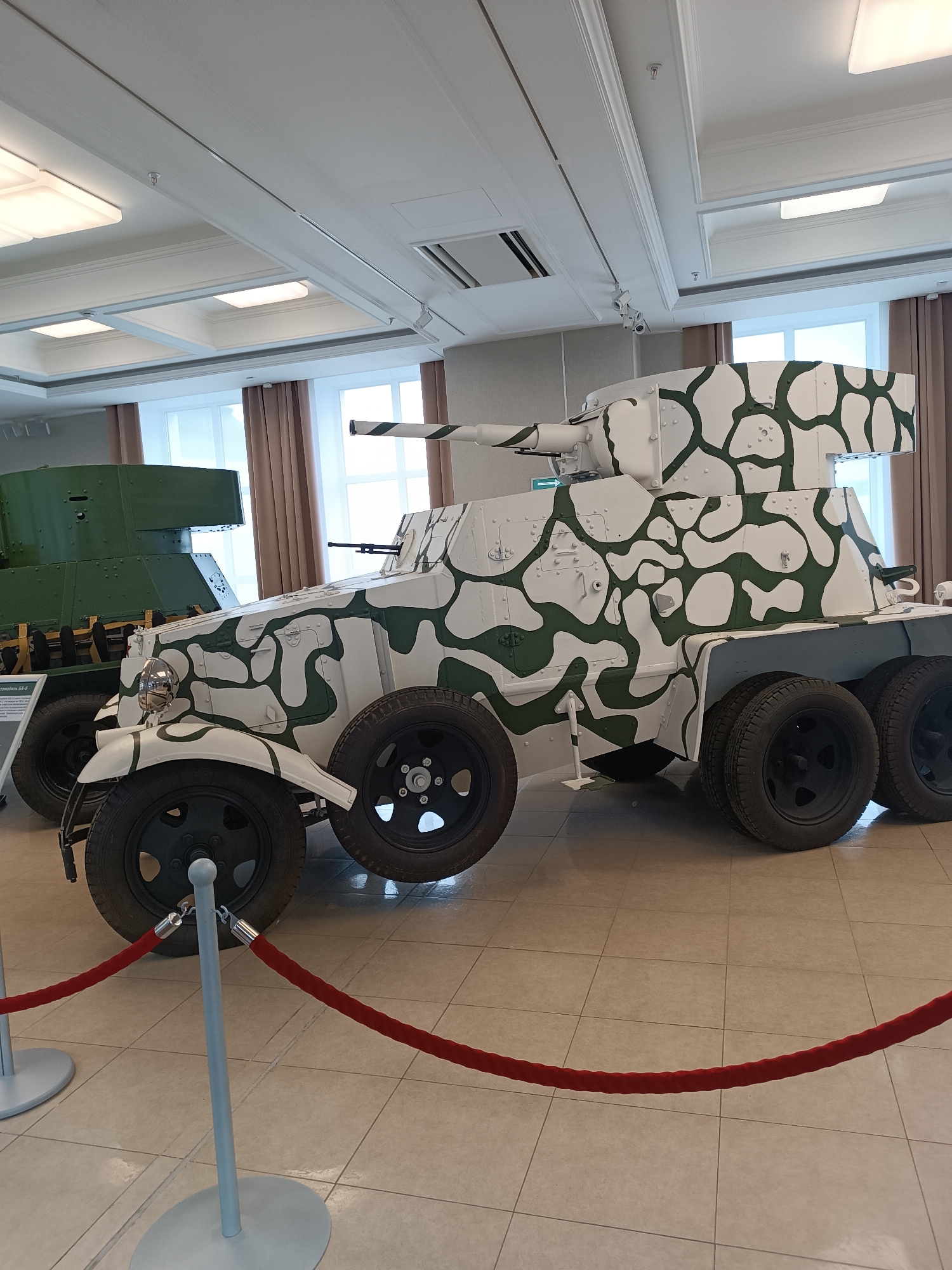 The image shows an indoor exhibition hall in the city of Verkhnyaya Pyshma, Russia. The focal point is a white armored car with green camouflage patterns, likely a museum exhibit. To the left of this vehicle, a portion of a green tank is visible, also appearing to be part of the exhibit. Red velvet ropes on stanchions are positioned in front of the armored car, serving as a barrier for visitors. The hall has a tiled floor, a white ceiling with recessed lighting fixtures, and windows with beige curtains in the background. The overall environment suggests a museum or historical display.