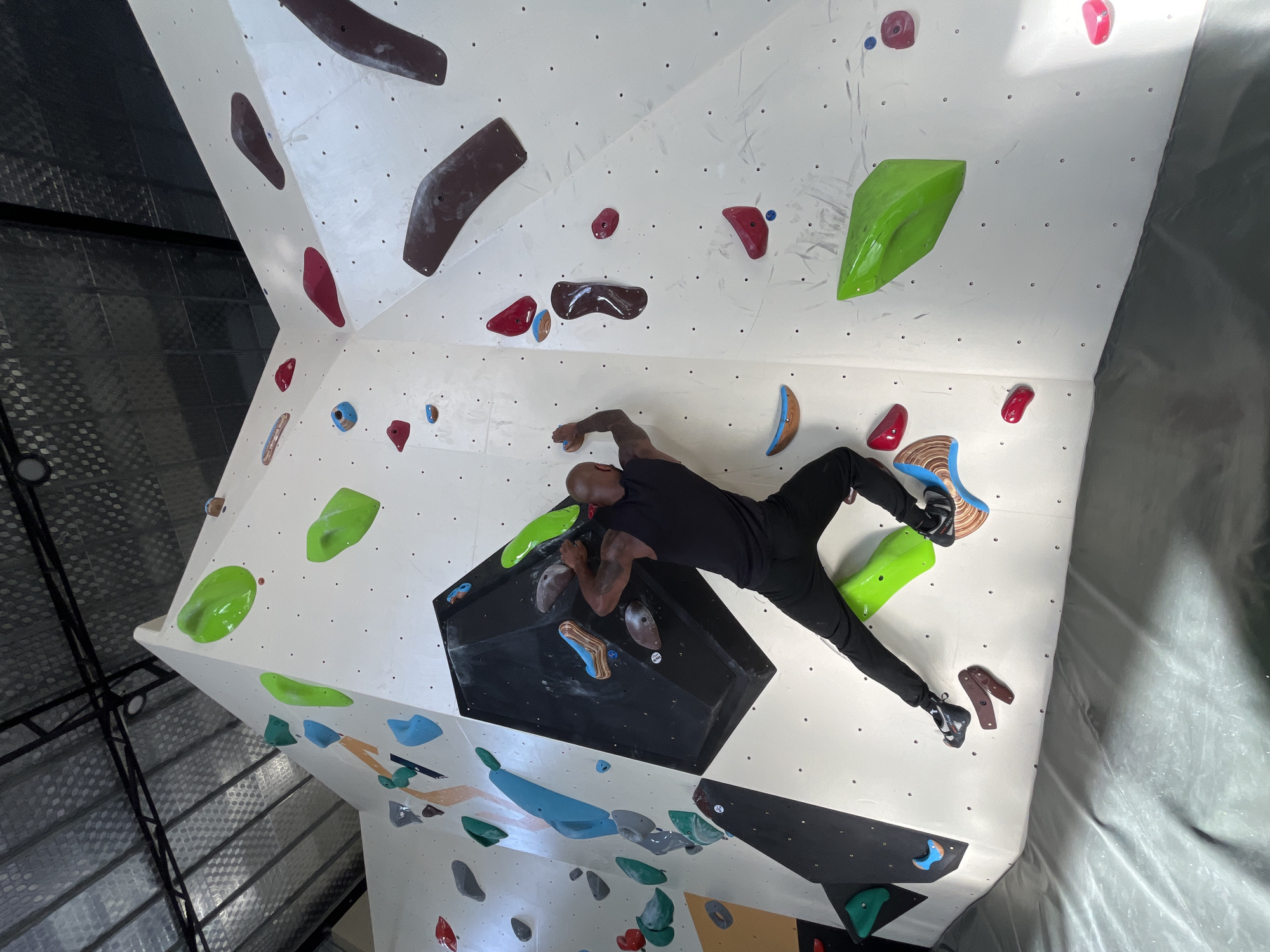 A male individual is observed bouldering on a white, multi-faceted climbing wall within an indoor facility in Đà Nẵng, Vietnam. The subject, a bald man with visible tattoos on his right arm, wears a black sleeveless top, black pants, and black climbing shoes. He is positioned on the wall, with his right hand gripping a dark, geometric volume and his left hand holding a brown and blue patterned climbing hold. His left foot rests on a large, bright green hold, and his right foot is placed on a smaller brown hold.
The bouldering wall is constructed with various angles and features several large black volumes. Numerous climbing holds of diverse shapes and sizes are affixed to the white surfaces, colored predominantly in green, red, blue, brown, grey, and orange. Below the wall, a continuous grey crash pad covers the floor, providing a safety surface. The ceiling above features an industrial design with a corrugated metal roof, black support beams, and exposed light fixtures.