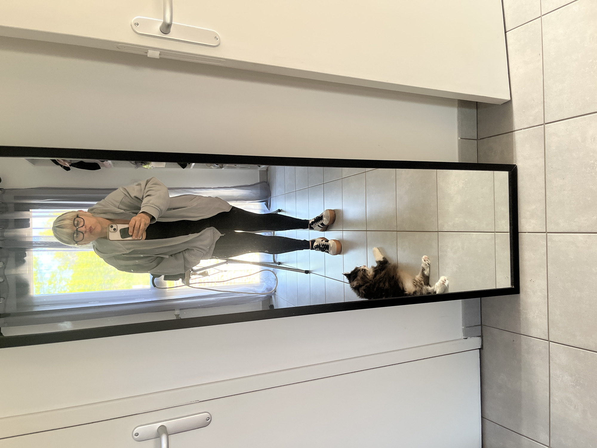The image shows a woman reflected in a full-length mirror in a tiled bathroom in Chambœuf, France. The woman is standing and taking a selfie with her phone. She is wearing glasses, a light grey hoodie, a black shirt, dark leggings, and sneakers with black stripes. A long-haired cat is lying on its back on the tiled floor in the reflection. The background behind the woman appears to be a window with outdoor lighting. The mirror is framed in black and is mounted on a white cabinet. The bathroom floor is covered in light grey tiles.