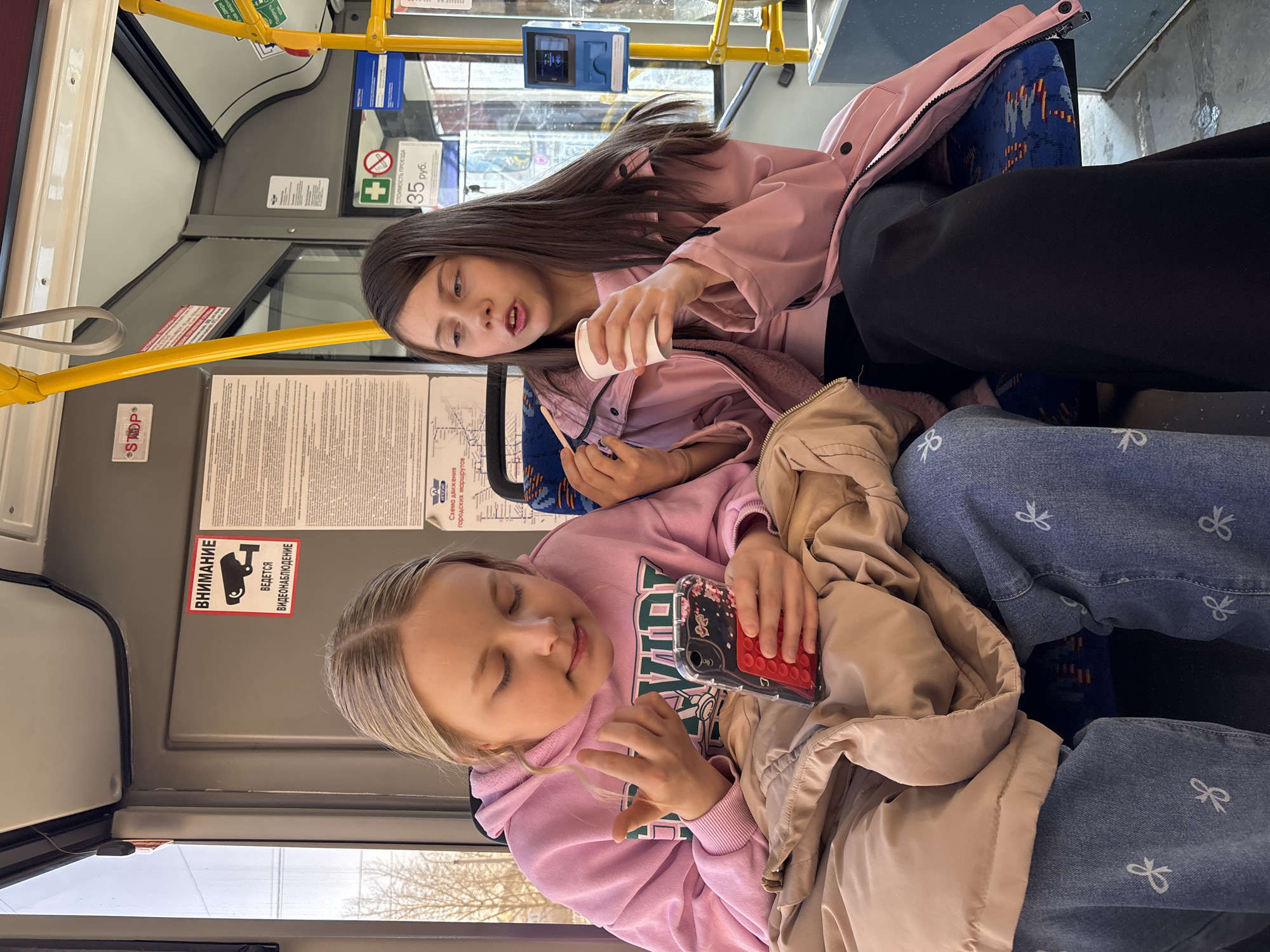 Two girls sit side-by-side on a bus. The girl on the left wears a pink hooded sweatshirt, light-blue jeans with white bow designs, and has a tan jacket covering part of her. She holds a red-cased mobile phone. The girl on the right wears a pink jacket, has long dark hair, and holds a small white paper cup with a straw. A sign on the bus shows a "35 руб" fare and camera icon indicating video surveillance, which is located in Izhevsk, Russia.