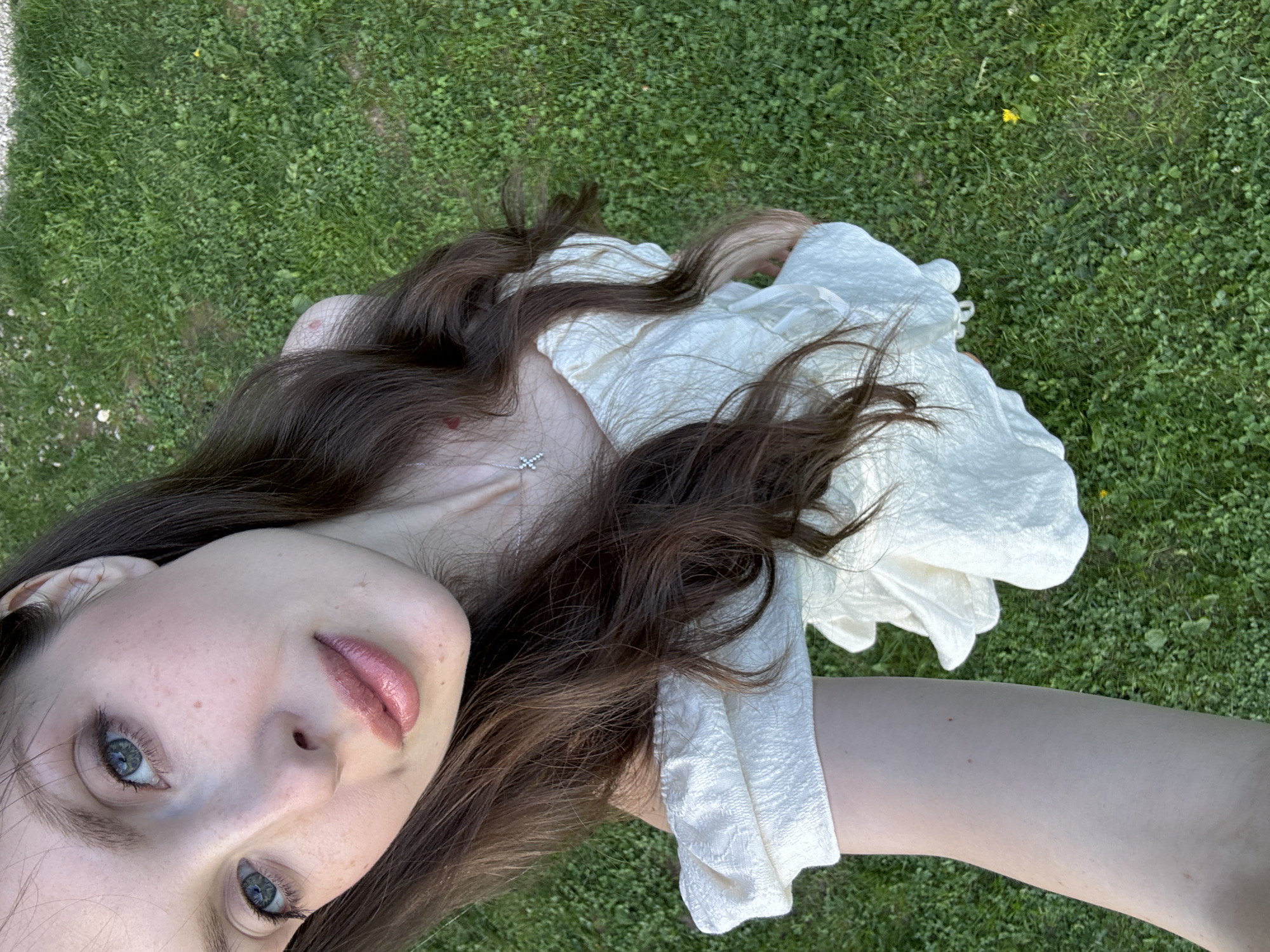 A young woman lies on a grassy area. Her long brown hair spreads around her, and she wears a white dress. She looks up into the camera with blue eyes. A silver necklace with a cross pendant is visible on her chest. The grass appears to be a lawn or field, and the lighting suggests it is daytime. The location is unknown, but the prompt suggests it may be in Prudki, Russia.