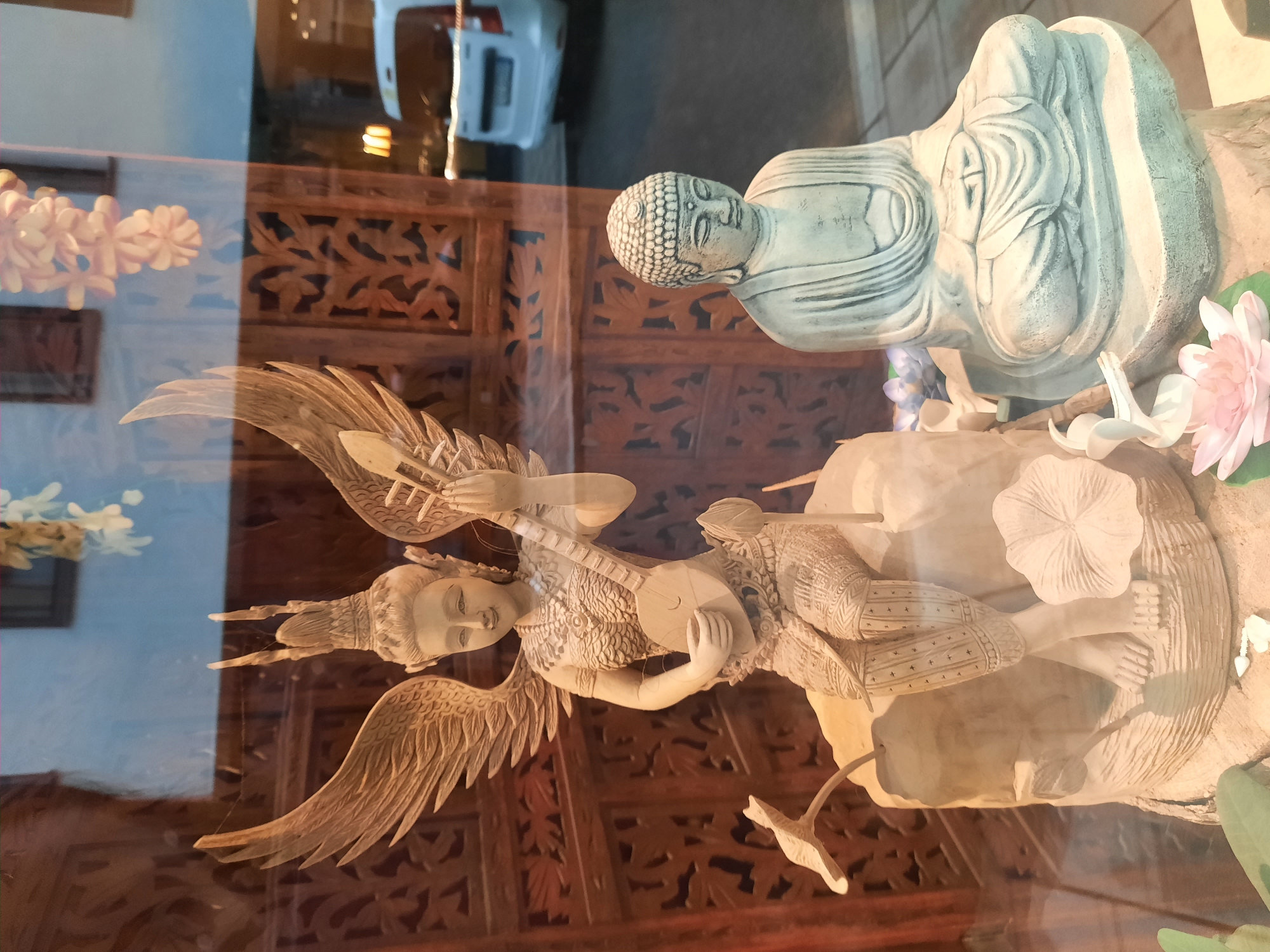 The image shows a store window display in Rosenheim, Germany, featuring two statues. The statue on the left is a light-colored, carved figure of a winged woman playing a stringed instrument, seated on a rock with lotus flowers. The statue on the right is a grey, seated Buddha figure. The background of the display includes carved wooden panels and reflections of the street outside, including a white vehicle. The lighting suggests it is daytime, possibly late afternoon or early evening, with a reflection of outdoor lights visible.