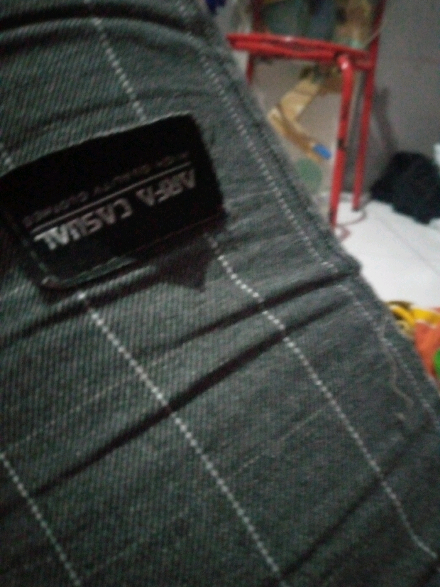 The image shows a close-up of a dark-colored, plaid fabric, likely clothing, with a black label attached. The label has white text that appears to read "ARFA CASUAL HIGH QUALITY CLOTHES". The background is blurred and indistinct, but appears to show a red metal structure and other objects. The lighting is dim. This scene is located in the city of Sumberwono, Indonesia.