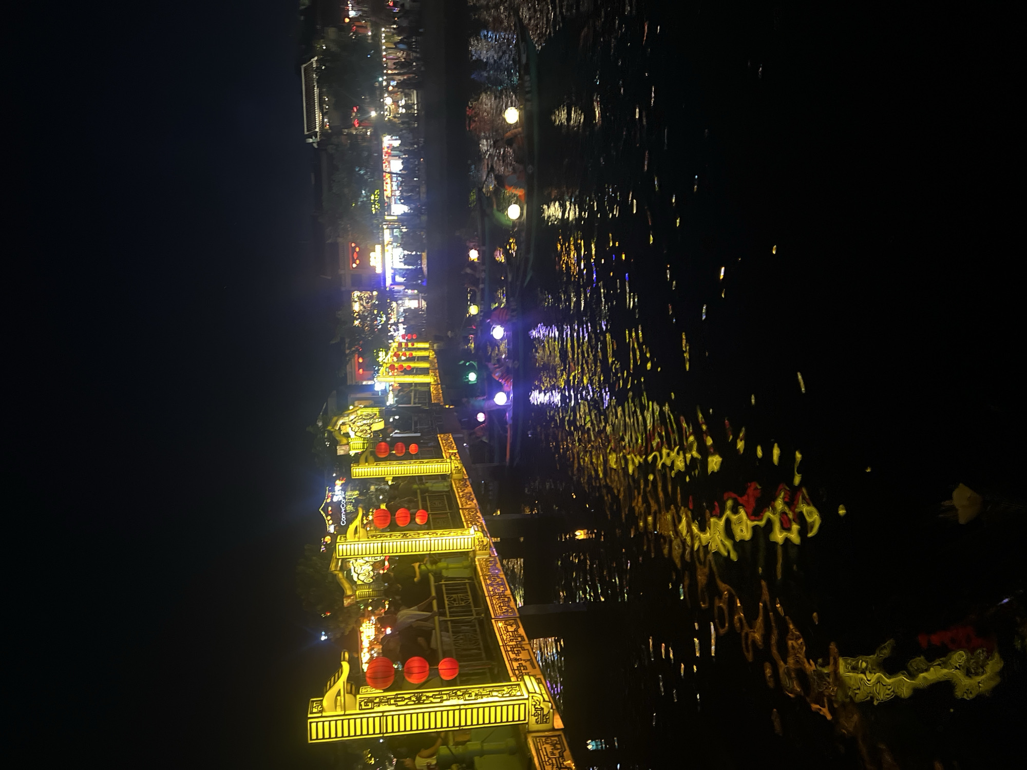 The image captures a nighttime view of the iconic Japanese Covered Bridge in Hoi An, Vietnam. The bridge is illuminated with vibrant yellow lights and adorned with traditional red lanterns, casting a warm glow. The water below reflects these lights, creating a shimmering effect. Several boats, also decorated with colorful lights, are navigating the water, with people visible inside them. The background shows the buildings and bustling streetscape of Hoi An, also illuminated and appearing lively. The overall scene depicts a festive and picturesque atmosphere characteristic of Hoi An at night.