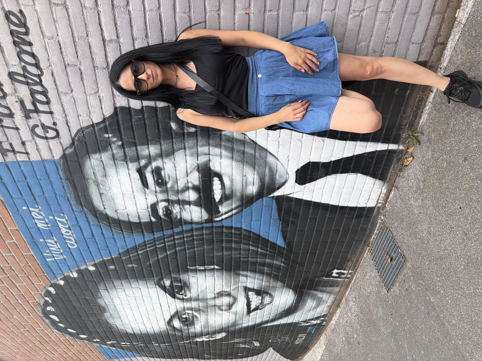 A woman with black hair, sunglasses, a black tank top, and a denim skirt leans against a brick wall adorned with a mural. The mural depicts two faces in black and white, with blue accents and white script. To the right of the mural, the name "G. Falcone" is visible in black lettering. The woman is positioned on the right side of the frame, facing forward with her left leg bent and propped against the wall. A dark strap bag is slung across her body. The ground in front of the wall is asphalt with a metal grate. The mural is located in the city of Corsico, Italy, specifically at Via G. Falcone, 20094 Corsico MI, Italy, commemorating the magistrate Giovanni Falcone.