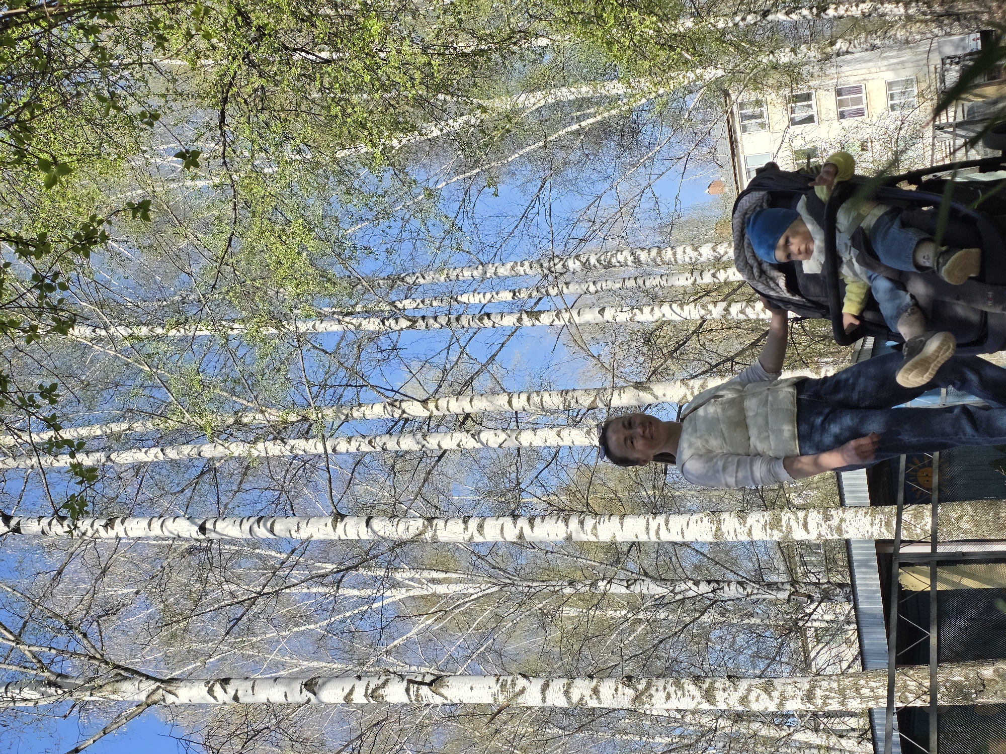 A woman in jeans and a white puffer vest stands behind a stroller with a child in it, next to birch trees with new leaves. The location is Chaikovsky, Russia. A building is visible in the background. A metal fence or enclosure is also visible.