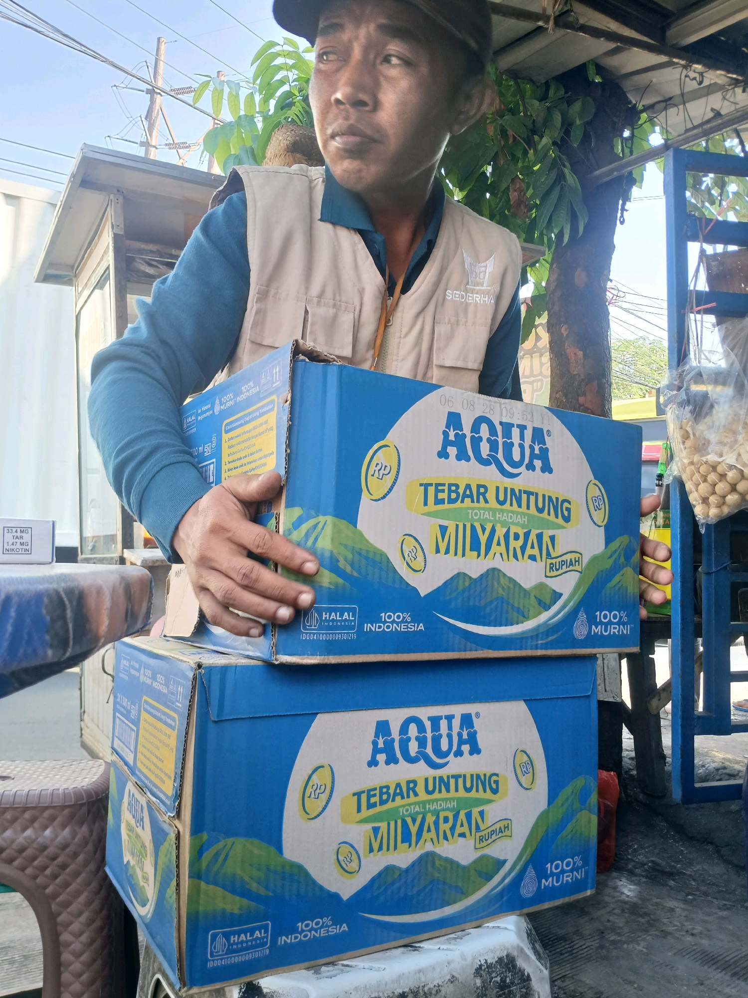 A medium shot captures a man in a tan vest over a blue long-sleeved shirt, holding two stacked blue cardboard boxes of AQUA bottled water. The boxes are decorated with green mountain imagery and text that includes "TEBAR UNTUNG MILYARAN" and "100% INDONESIA". The man wears a dark baseball cap and looks slightly upward and to his right. The background suggests an outdoor market or street scene in Bandung City, Indonesia, with overhead wires, a building structure, a tree with green leaves, and a blue metal frame. A stool is visible to the left, and a plastic bag filled with round, light-colored objects hangs to the right. The overall lighting indicates it is daytime.