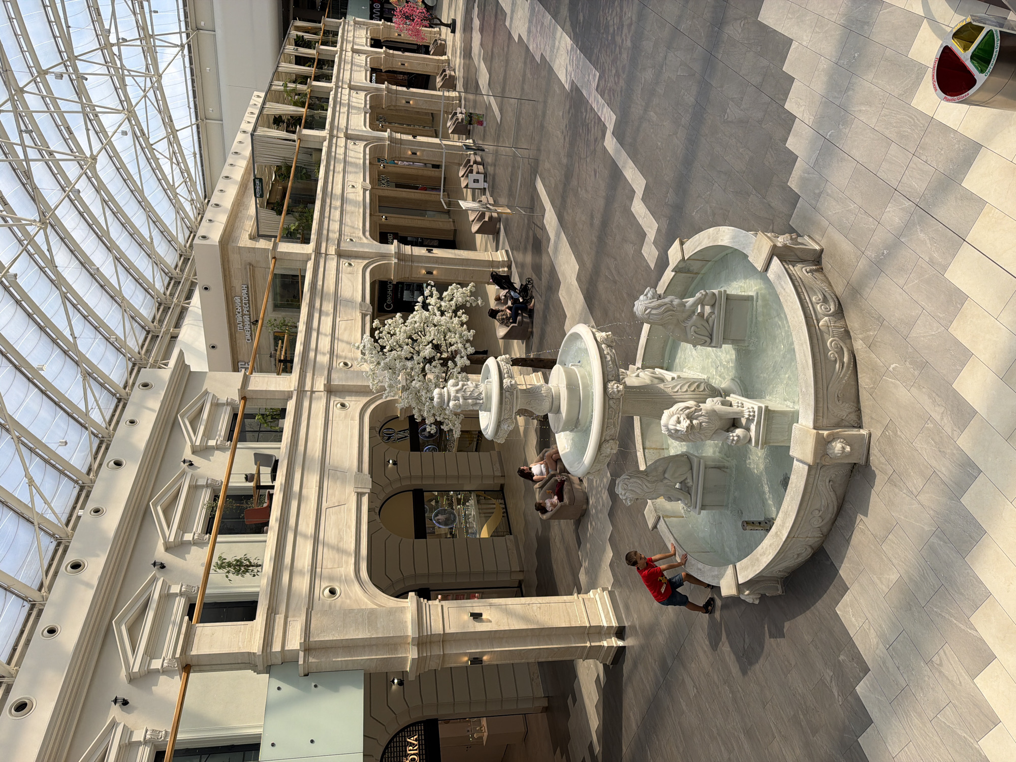 This is an aerial shot of the Atrium of the Ocean Plaza mall in Kyiv, Ukraine. The mall's architecture features a grand, light-filled interior with a glass roof, elegant arches, and cream-colored stone facades. In the center of the atrium, a multi-tiered white stone fountain with lion statues is prominently displayed. A young boy in a red shirt and blue shorts is leaning over the edge of the fountain, seemingly interacting with it. Several people are seated in lounge chairs in the background, enjoying the mall's ambiance. The floor is tiled with a pattern of light and dark gray squares, and shops line the upper levels of the atrium.