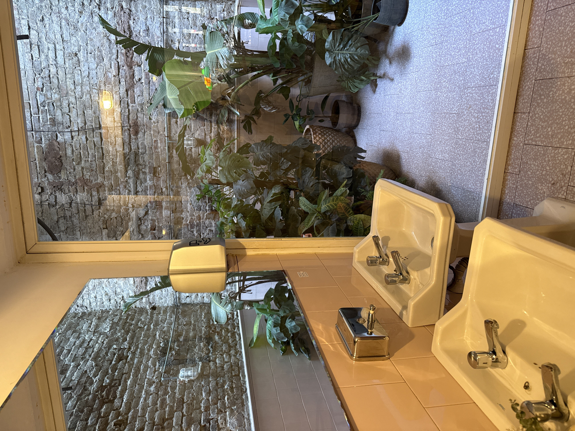 The image depicts a bathroom scene in what appears to be a building in Barcelona, Spain. The view through a large window reveals a brick wall covered in greenery, possibly an exterior courtyard or adjacent garden. The interior bathroom features two side-by-side white porcelain sinks with chrome fixtures. A chrome soap dispenser sits on the pink tiled countertop between the sinks. A mirror reflects the brick wall and plant life. The floor is tiled in a light beige speckled pattern, while the countertop has pink tiles. A hand soap dispenser is mounted on the wall above the counter.