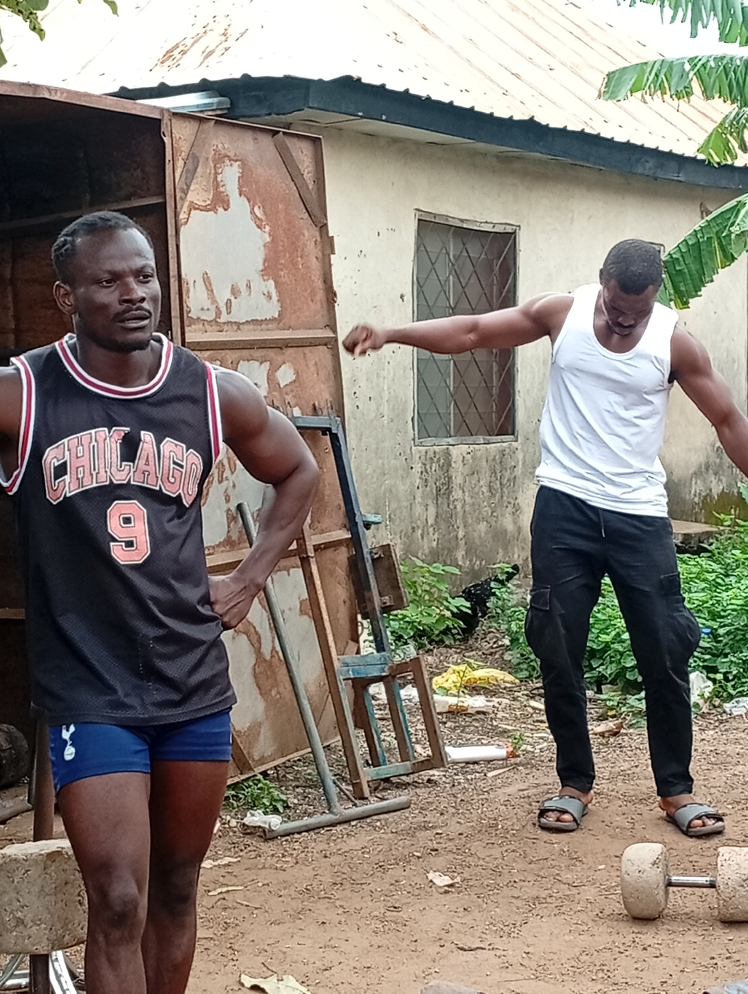 This outdoor image, taken in Akwanga, Nigeria, features two muscular men on a dirt ground, seemingly engaged in or resting from a workout.
On the left, a man with dark skin and braided hair wears a black mesh basketball jersey with "CHICAGO" and the number "9" prominently displayed in red and white. He has his hands on his hips and looks forward with a focused, serious expression. He is wearing blue athletic shorts with a small white logo, which appears to be the Tottenham Hotspur crest, on the left thigh.
On the right, another man with dark skin wears a plain white tank top and black cargo pants. His head is bowed, and his arms are extended outwards, suggesting he might be stretching or posing after exercise. He wears grey slide sandals.
In the foreground, to the right, are two concrete dumbbells resting on the dirt, indicating a makeshift workout area. The background includes a simple, light-colored building with a corrugated roof, and a rusted metal structure, possibly a door or gate. Various green foliage, including broad banana leaves, adds to the outdoor, somewhat rustic setting.
The lighting suggests it is daytime, likely overcast, given the soft shadows. The overall scene depicts a dedication to physical fitness within a modest, outdoor environment.